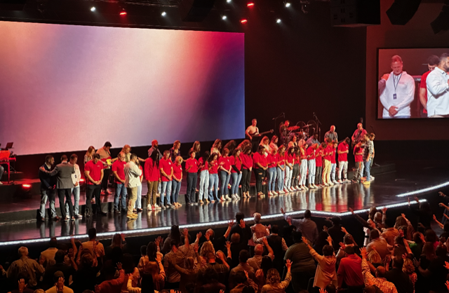 Large group of people in red shirts on a stage with a big screen, while an audience applauds.
