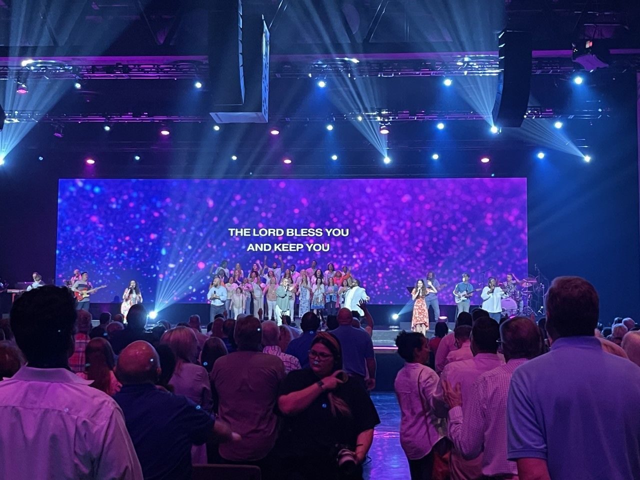 People attending a worship service; large stage with musicians and choir under purple lights; text: