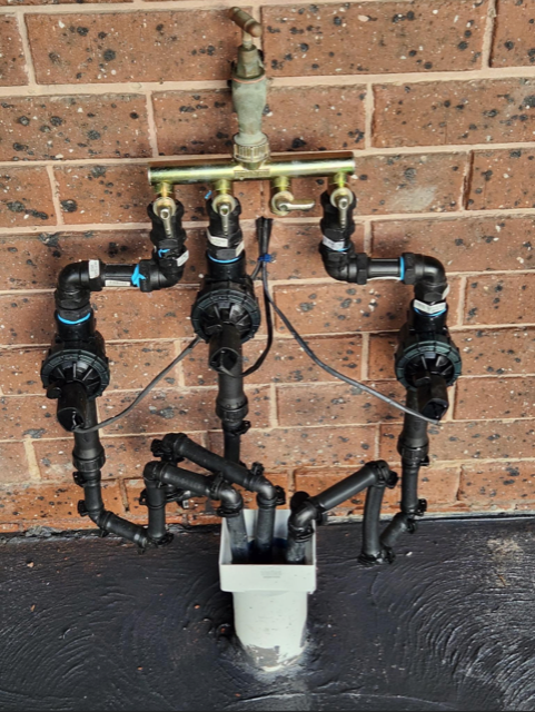 Black plumbing manifold with multiple valves and pipes mounted on a brick wall, ending in a white bucket below