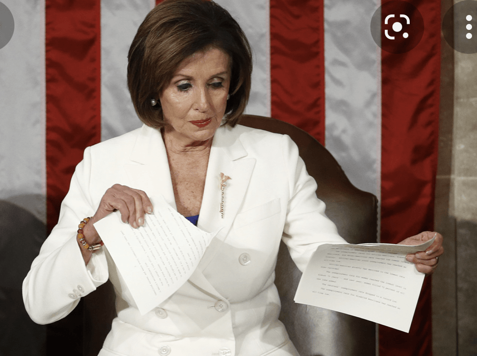 Nancy Pelosi tearing papers, seated in front of an American flag backdrop. She wears a white suit, focused expression.