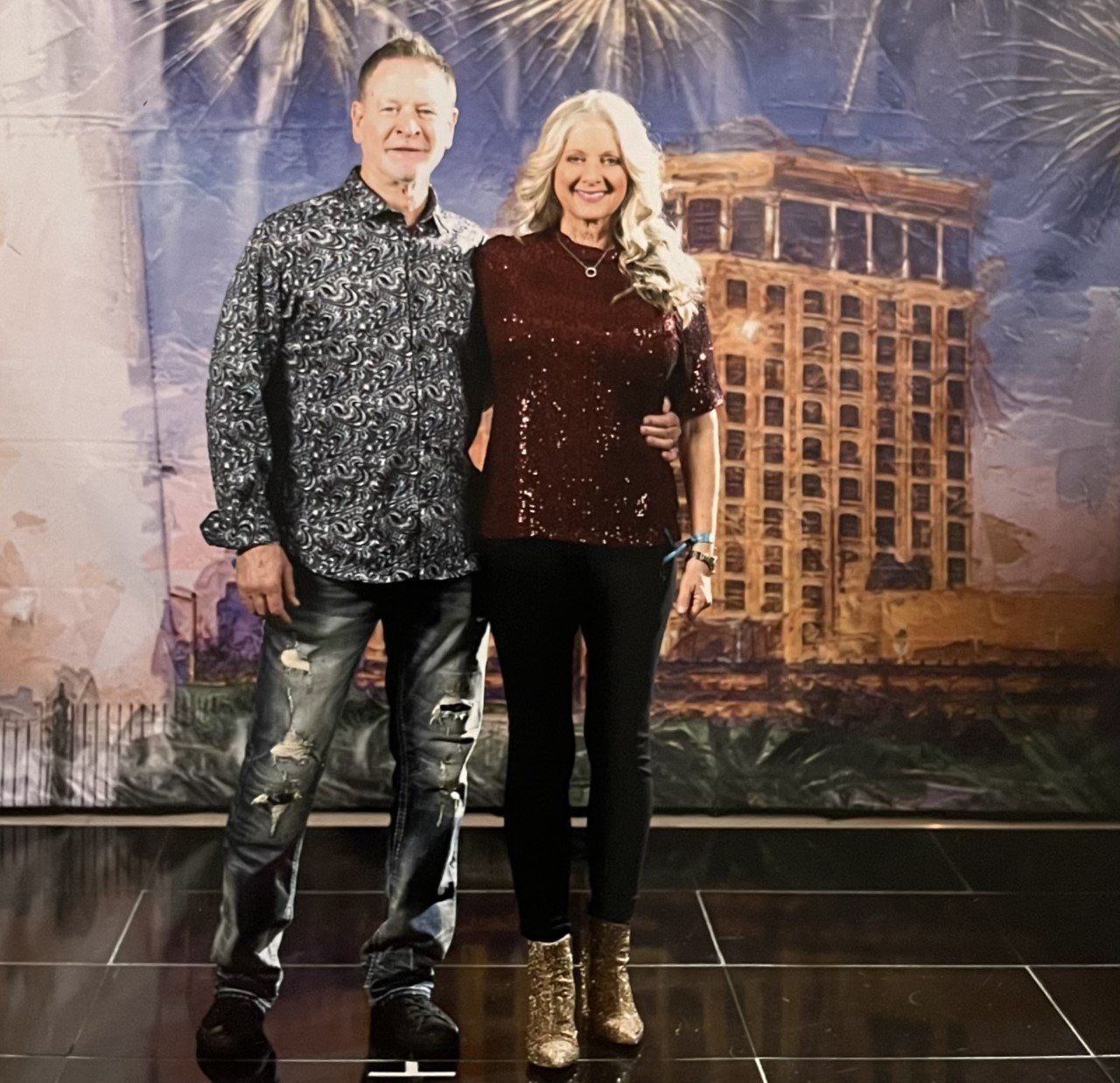 Couple poses in front of a colorful building with fireworks. Man wears printed shirt and jeans, woman in sparkly top and black pants.