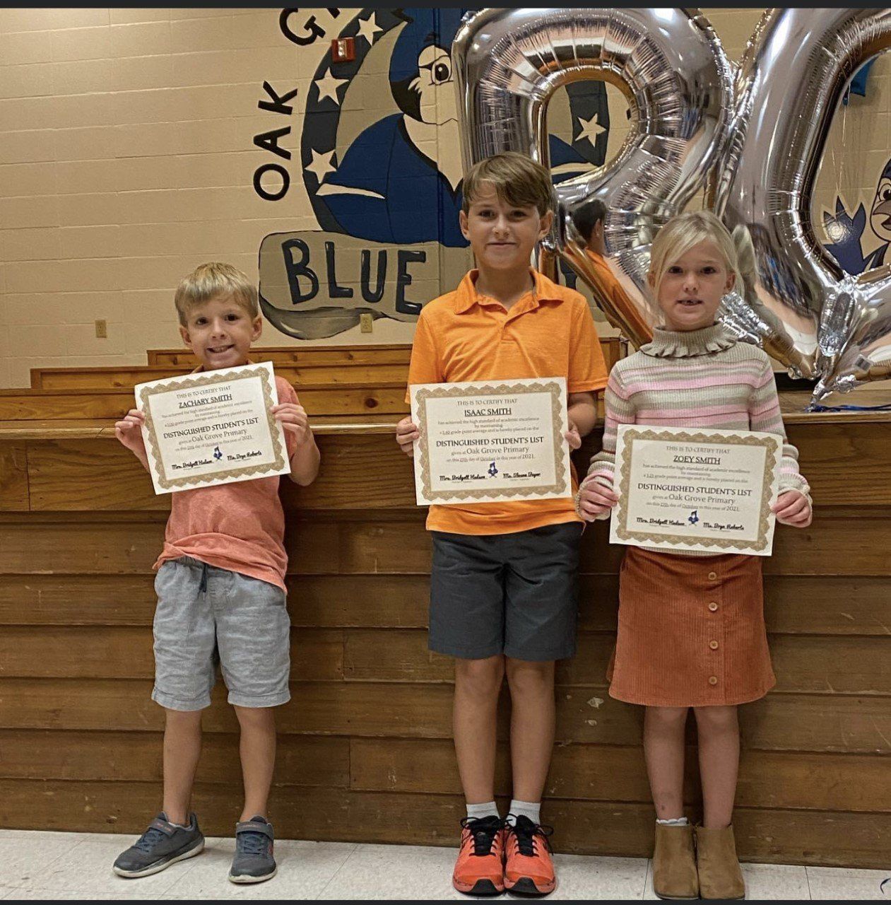 Three children holding certificates on a stage. A blue and white school sign and balloons are in the background.