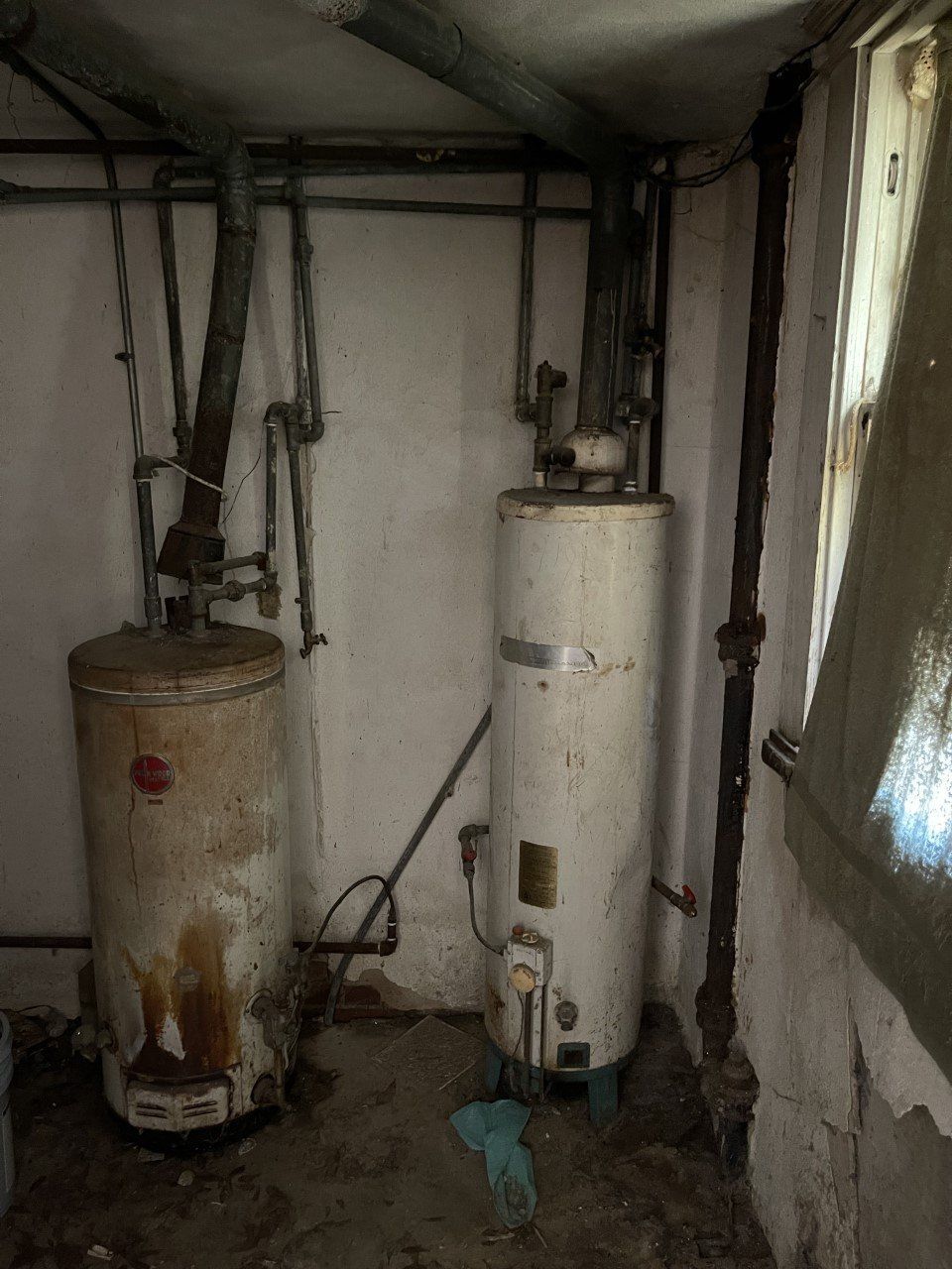 Two old, rusty water heaters in a dingy basement corner. One tank shows water stains.