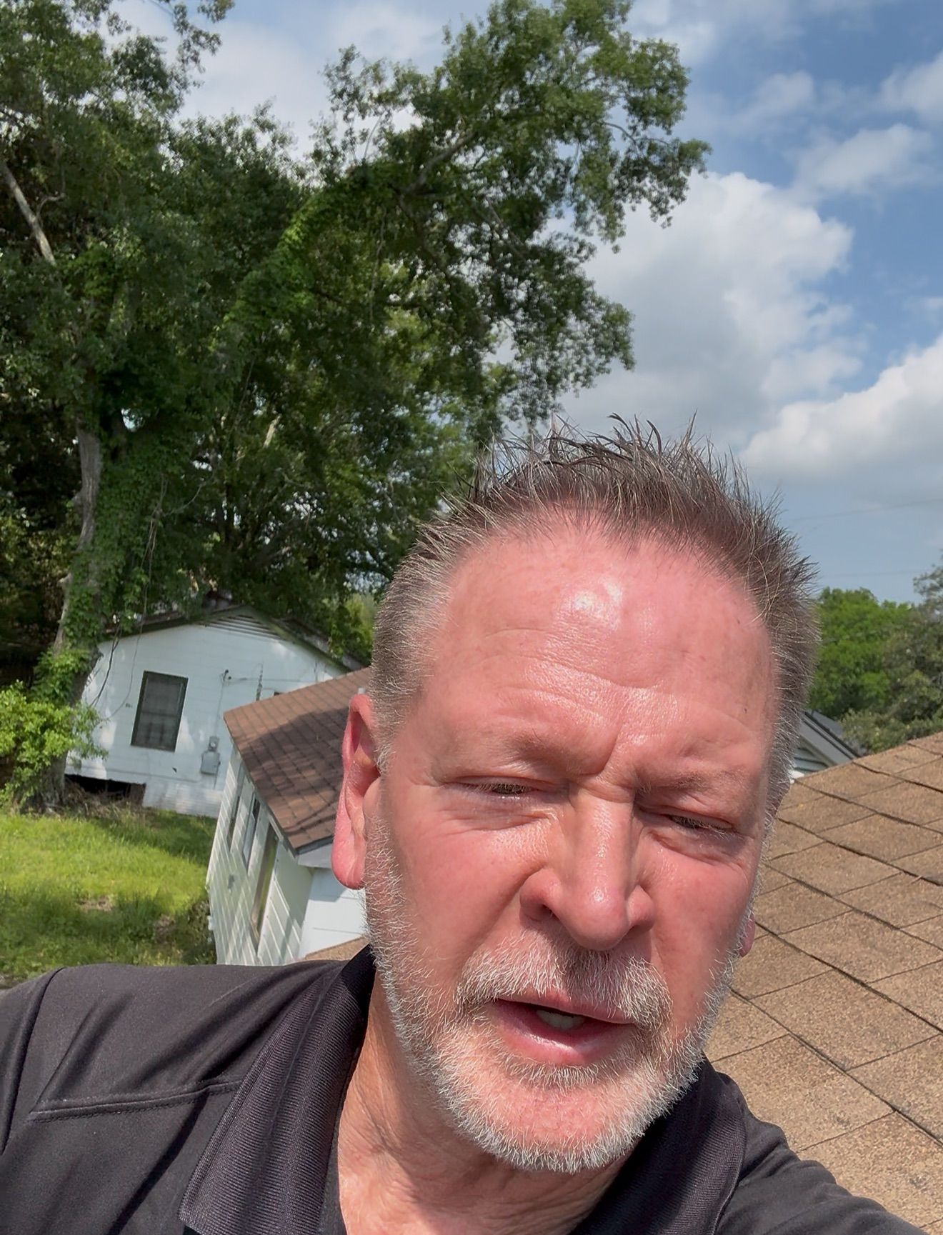 Man taking a selfie outdoors beside a house and trees under a cloudy sky