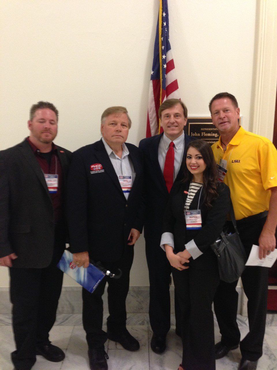Five people standing by a US flag. Men in suits and a yellow shirt, a woman in a black outfit. Indoors, formal setting.