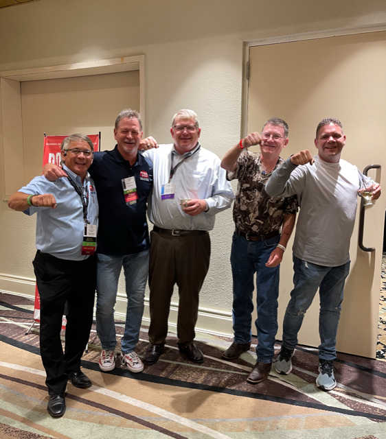 Five men smiling, posing with fists up in a hallway, some wearing conference badges.