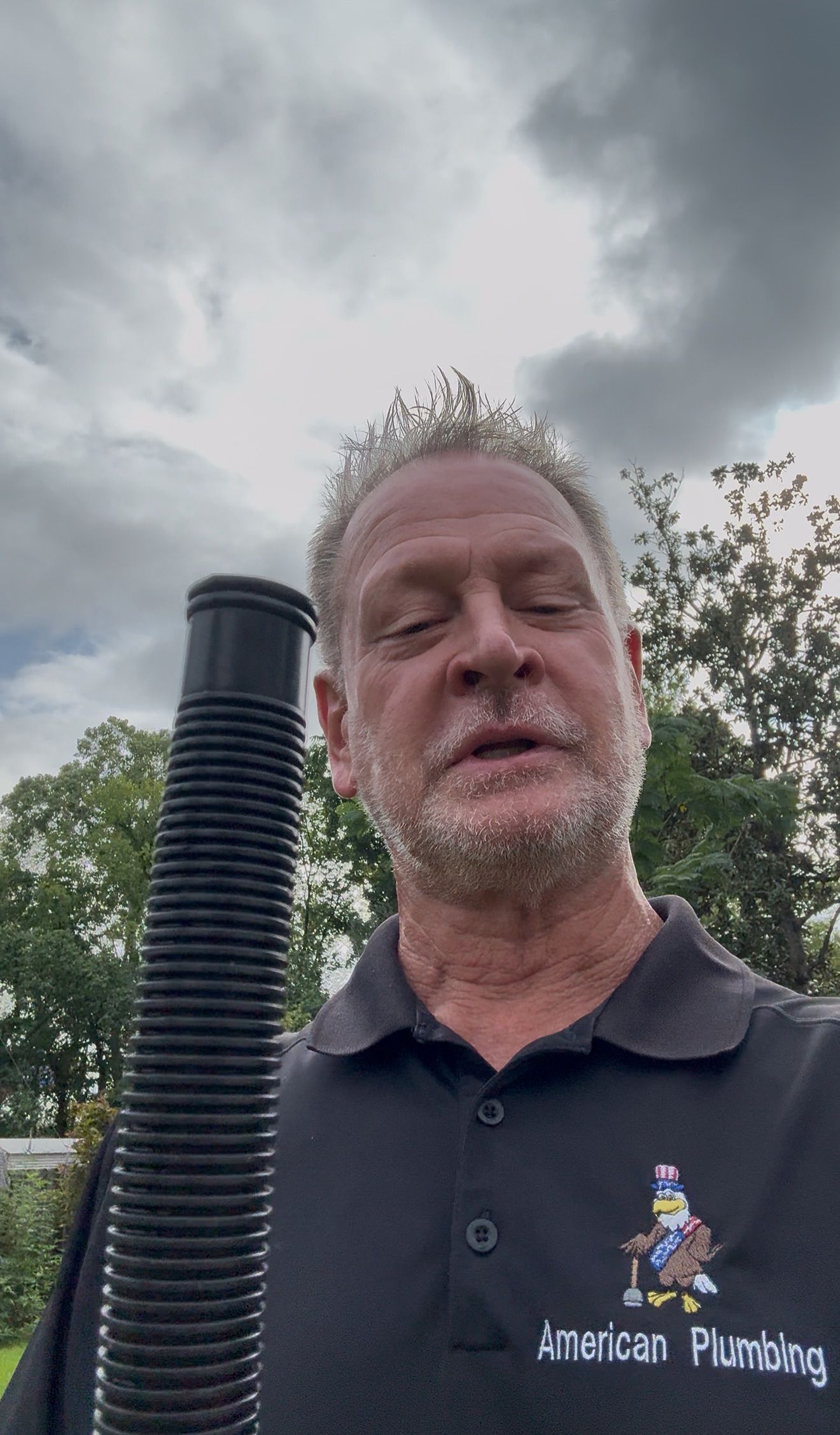 Man in a black polo stands outdoors beside a black vent pipe under a cloudy sky.