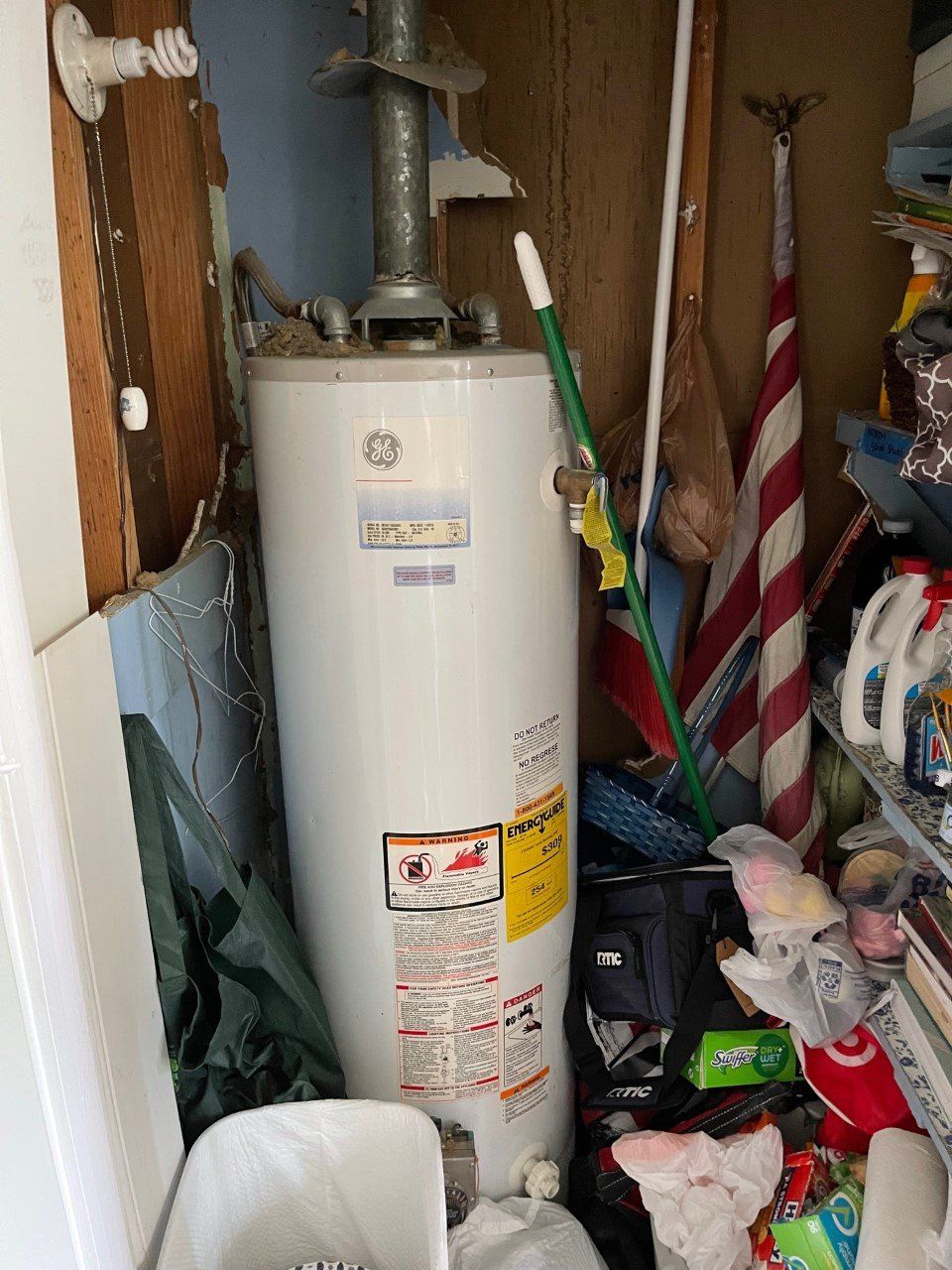 Water heater in a cluttered closet with cleaning supplies and tools around it.