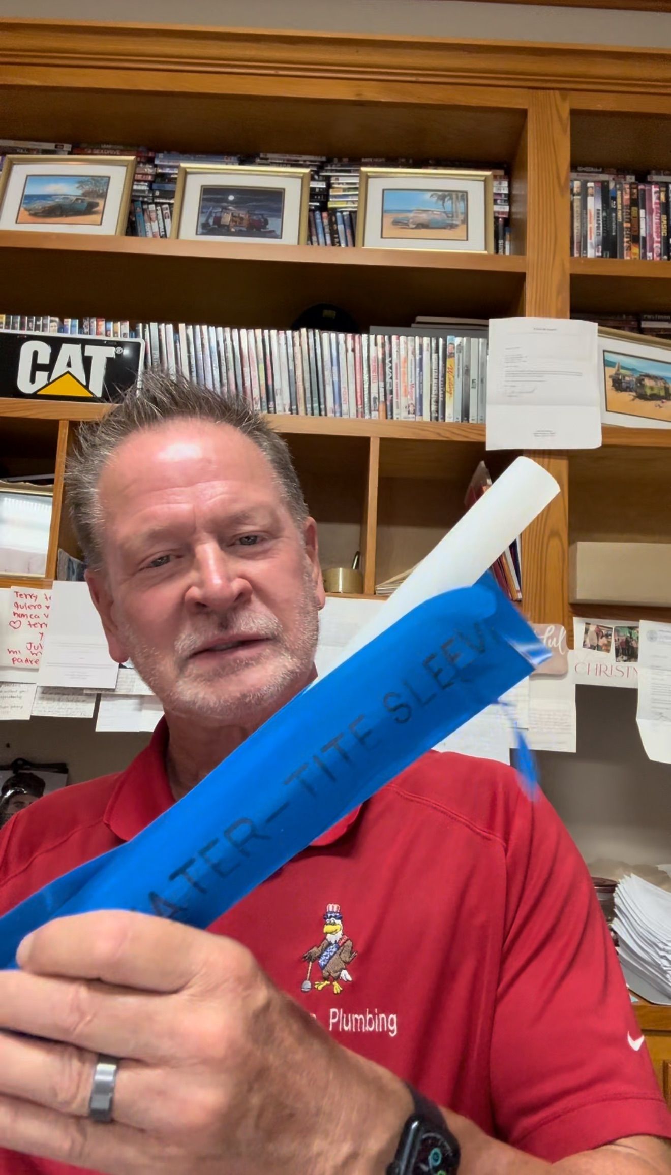 Man in red shirt holding a blue rolled object in a room with shelves of books and boxes