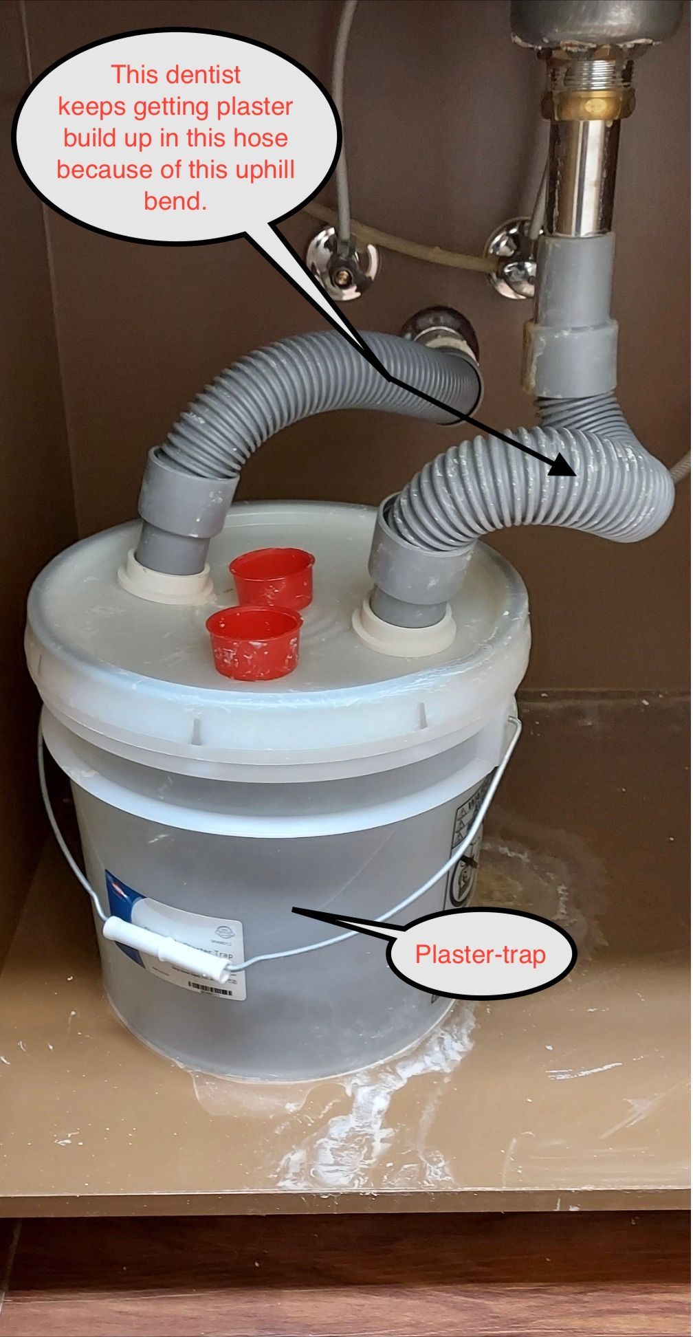 Gray bucket-style sump pump with white lid, red caps, and hoses connected to plumbing in a flooded area
