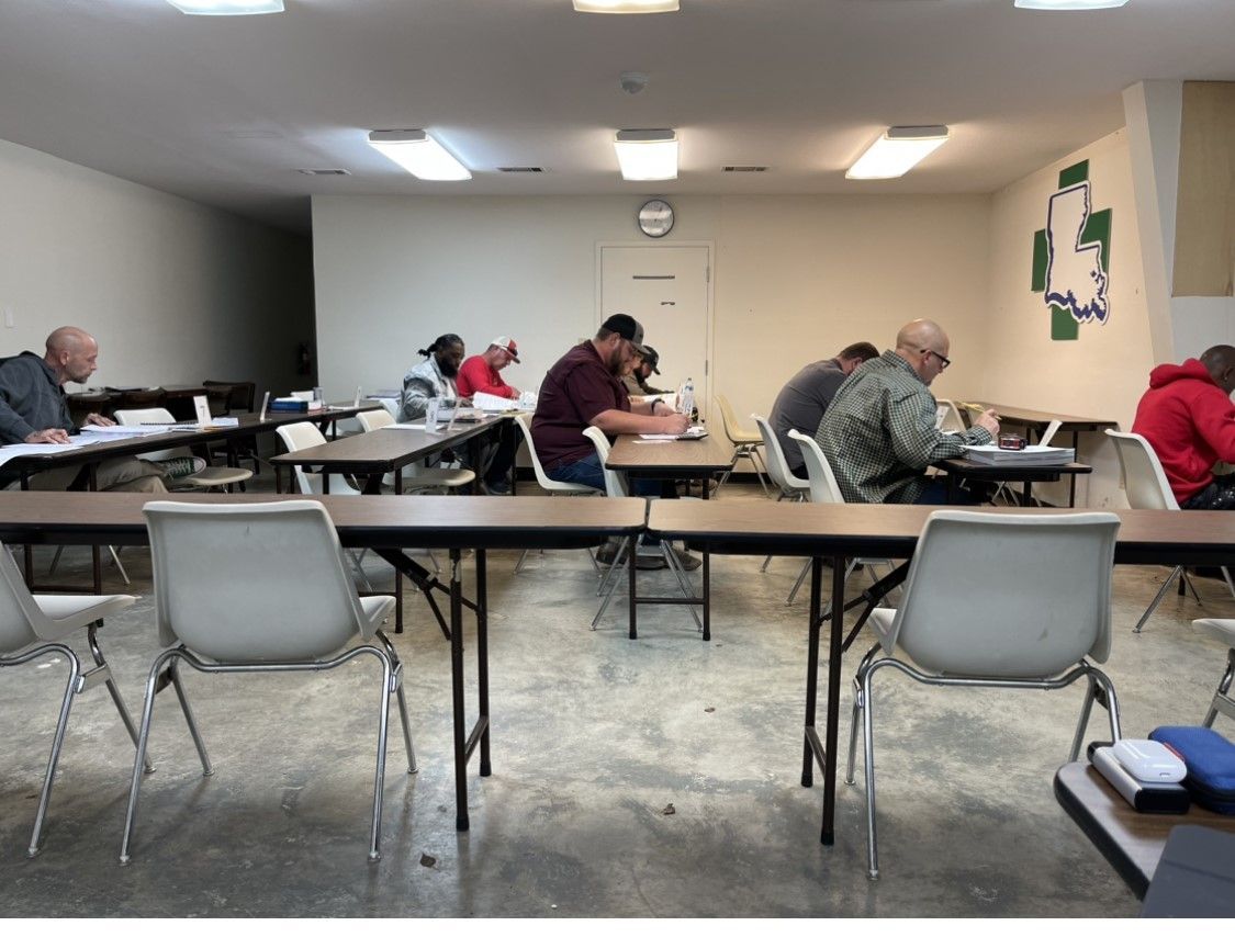 People seated at tables in a classroom, taking notes. White chairs, fluorescent lights, and a clock on the wall.