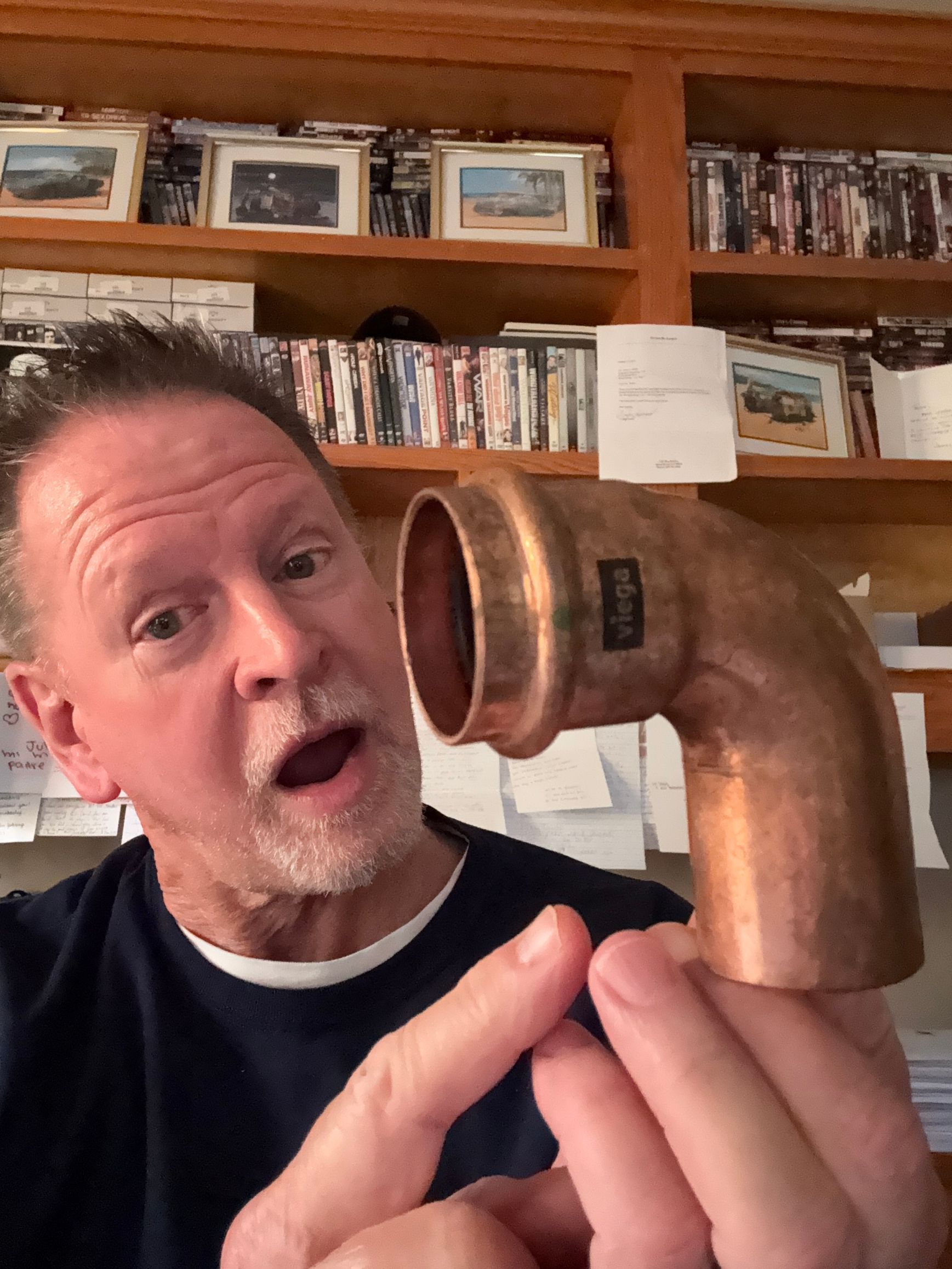 Man holding a copper pipe fitting close to his face in a workshop or office with shelves behind him