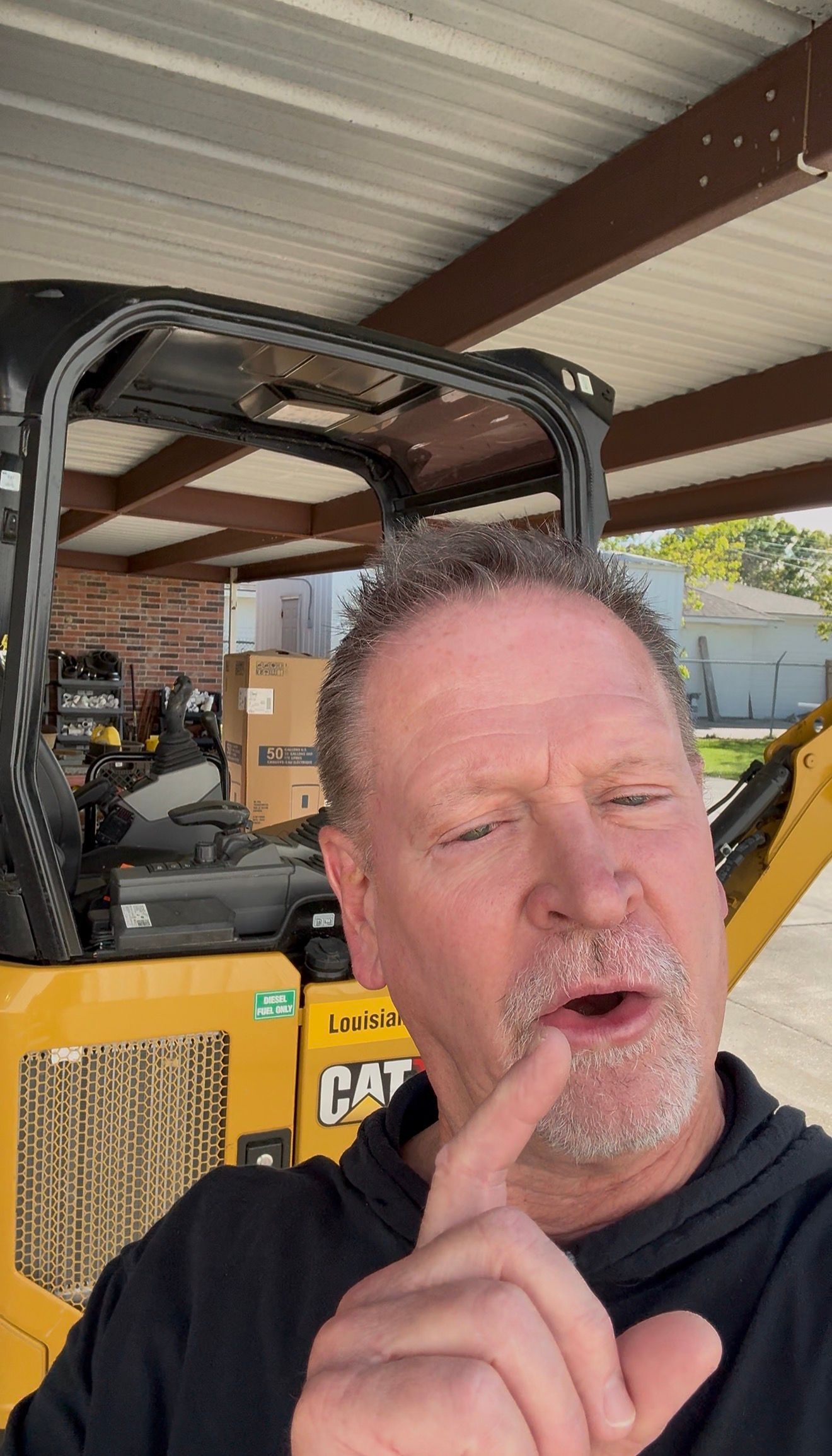 Man posing in a workshop, pointing upward beside a yellow forklift under a metal roof.