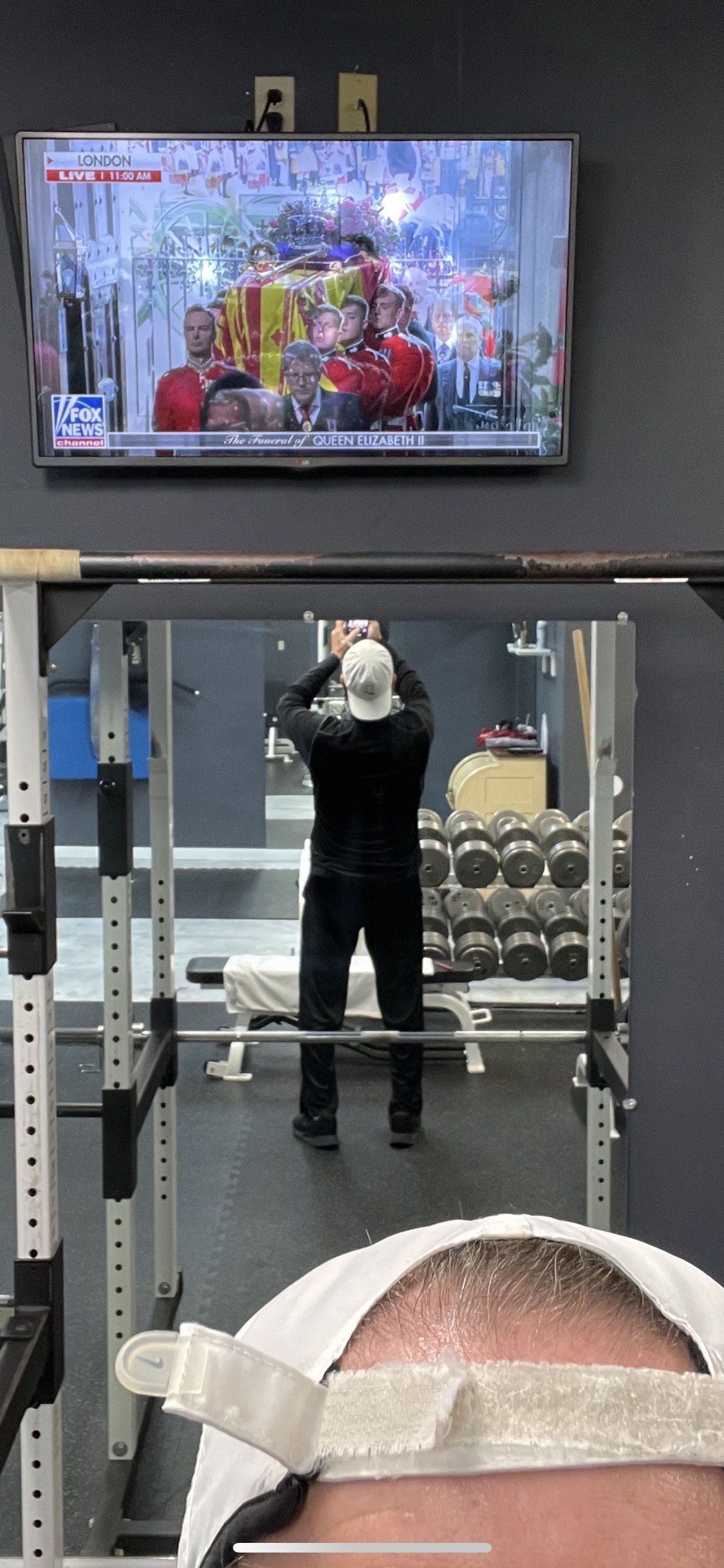 Gym mirror selfie with a person in dark clothes, weight machine, and TV screen above