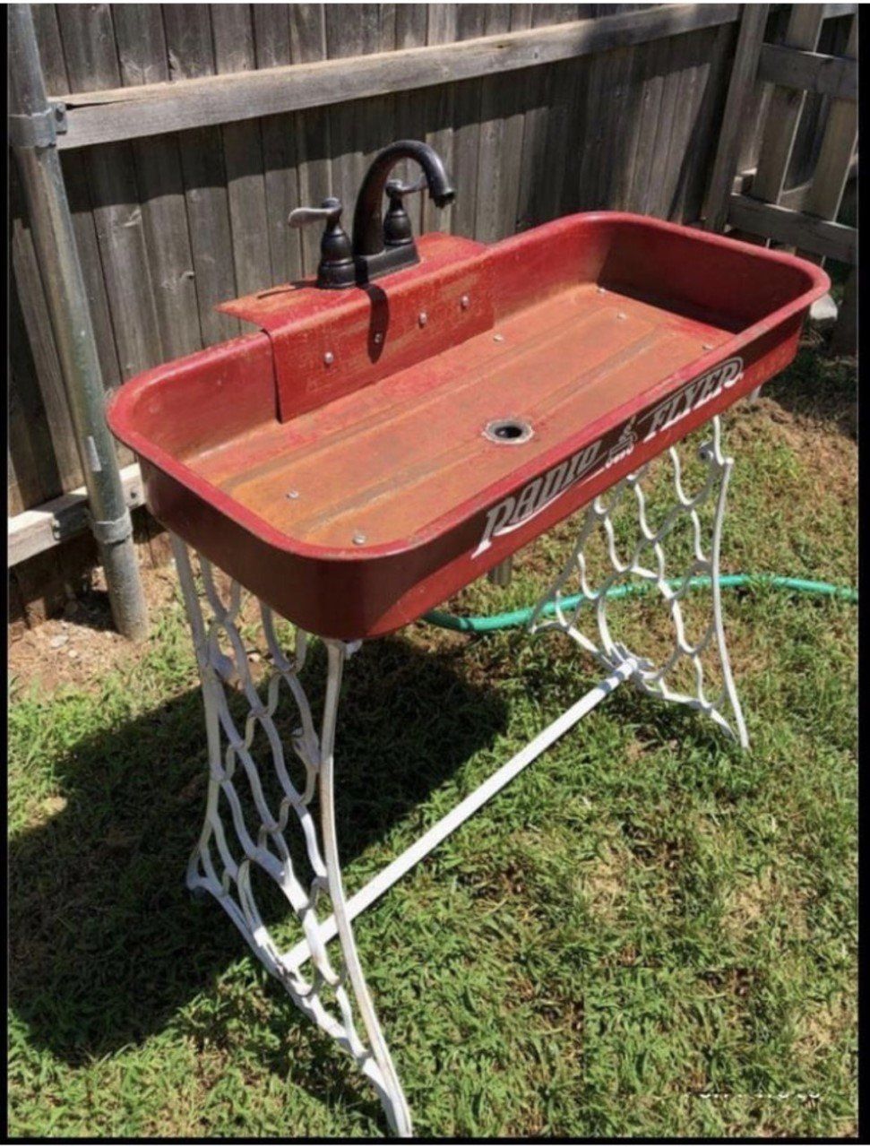 Outdoor sink made from a red wagon bed, brown wooden countertop, black faucet, and white decorative stand.