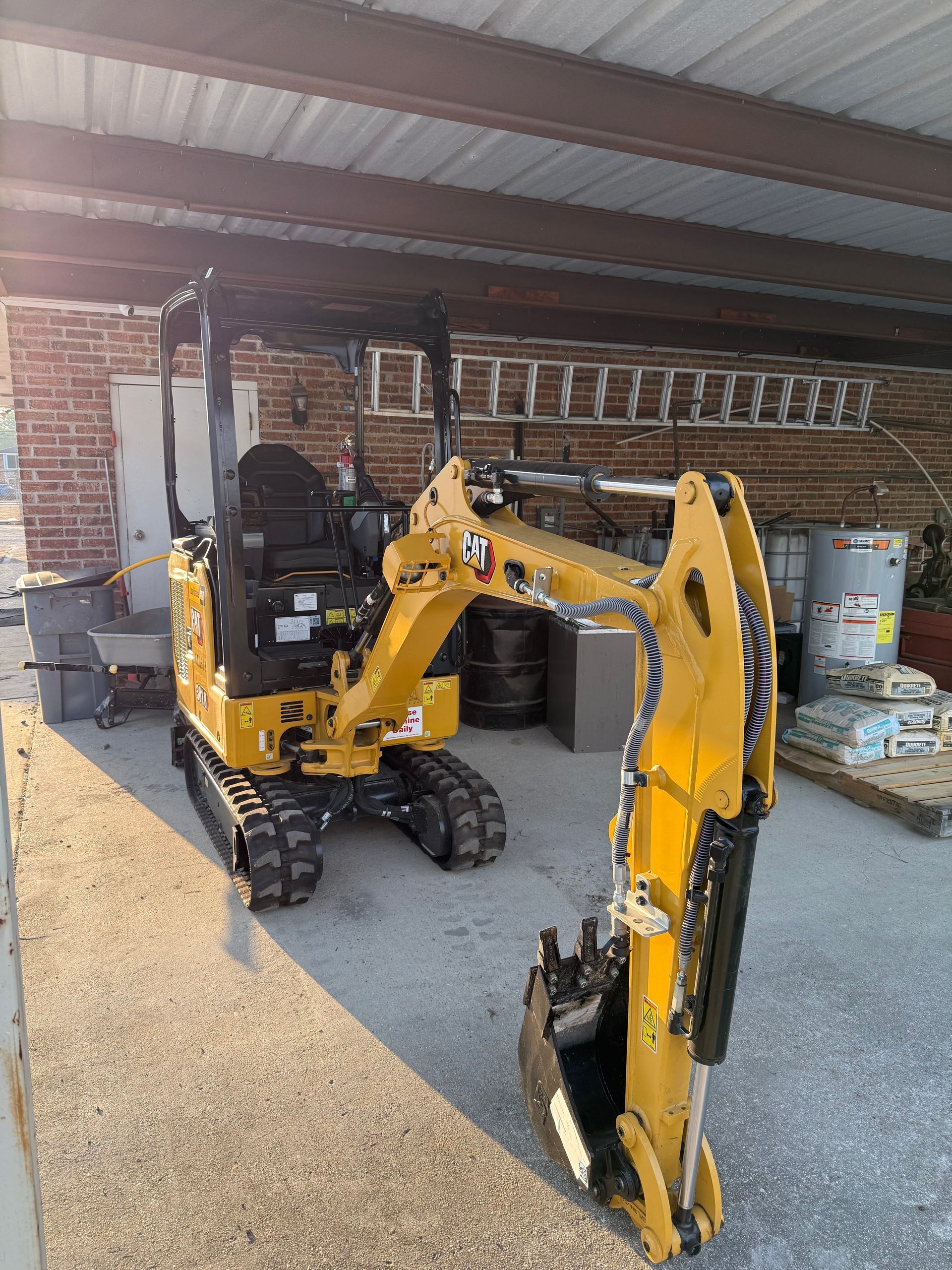 Yellow mini excavator in a gravel-covered garage beside a brick wall and stacked equipment