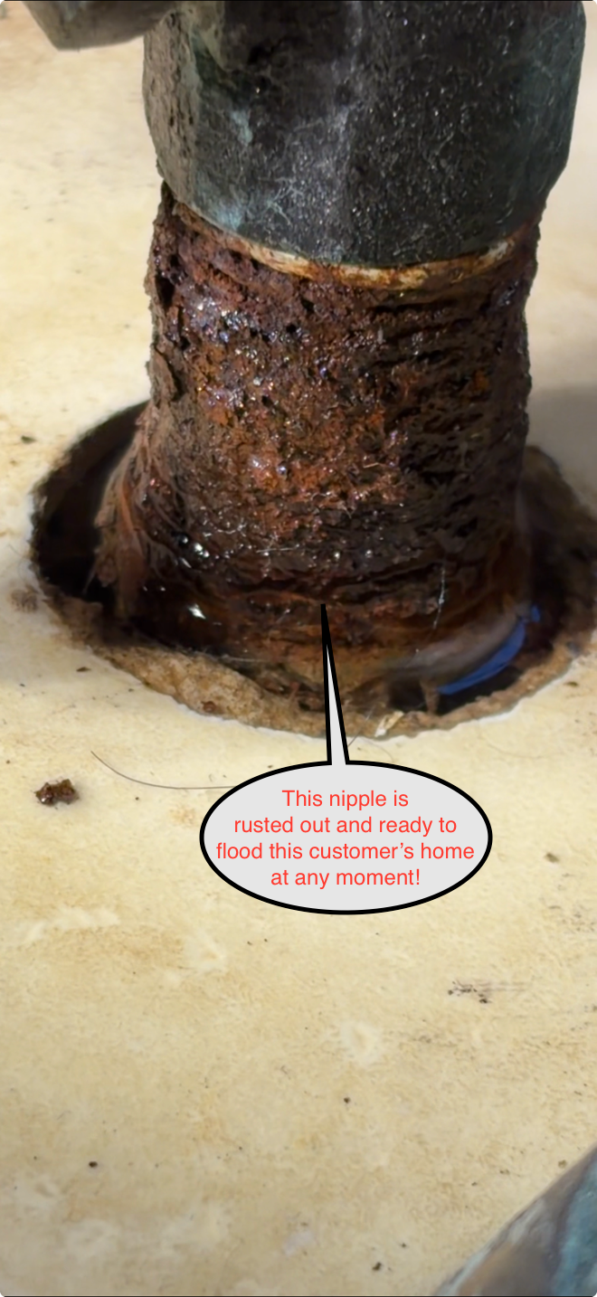Rusty corroded pipe joint on a light floor, with a small warning label below.