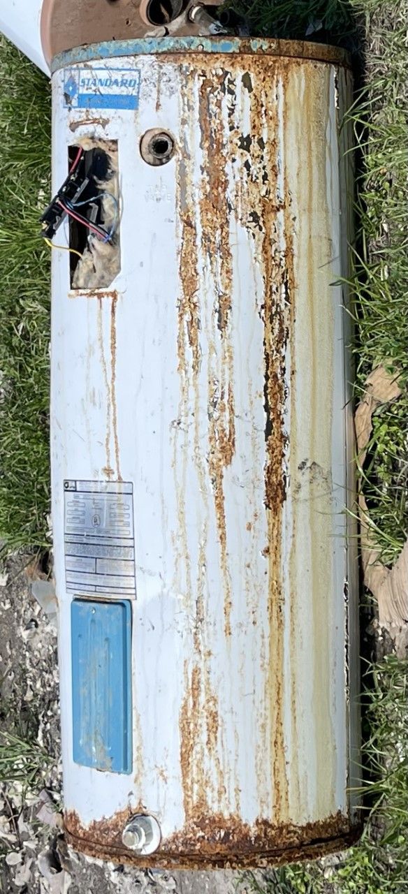 Rust-streaked white cylindrical tank standing outdoors beside grass and a wall.
