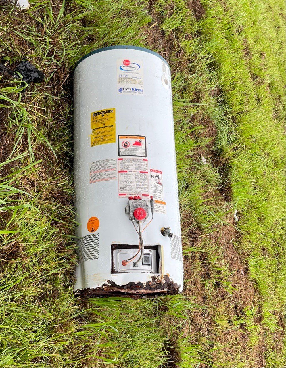 White cylindrical tank lying on grass, with warning labels and a red valve near the bottom.