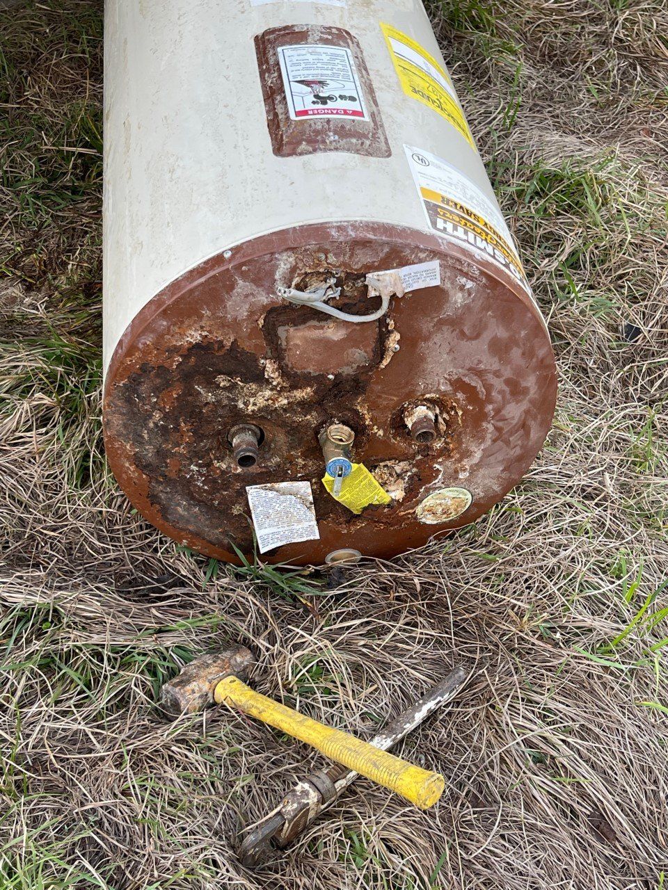 Rusty metal tank end on grass with a yellow-handled tool and bolts nearby