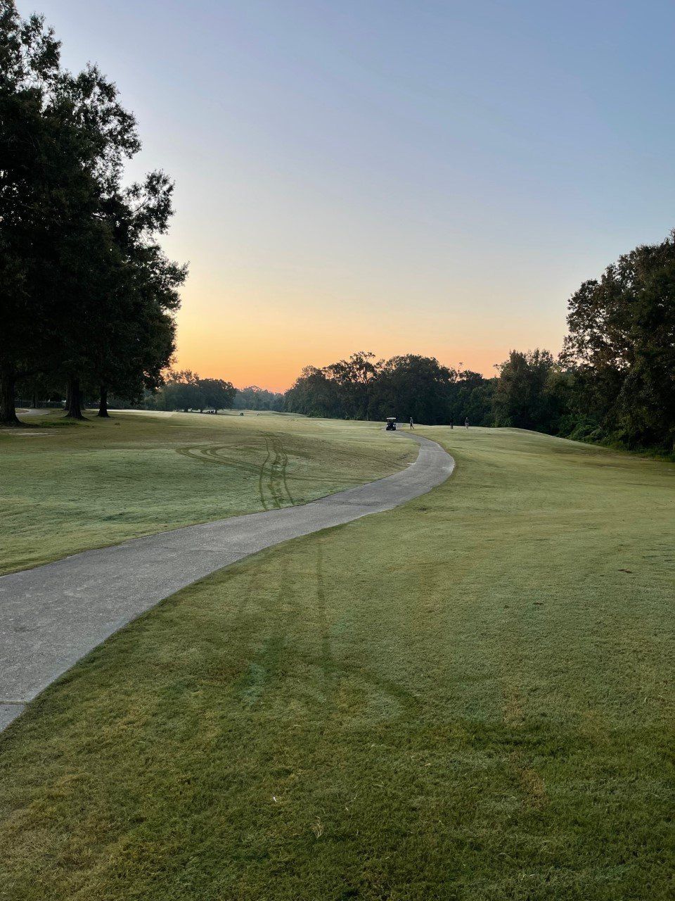 A paved path winds through a misty golf course at dawn, with trees lining the edges and a colorful sky.