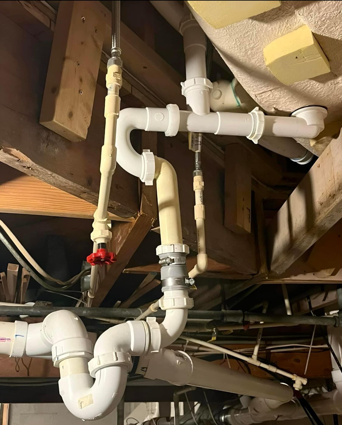 Exposed white PVC plumbing pipes and valves under a wooden floor joist in a basement