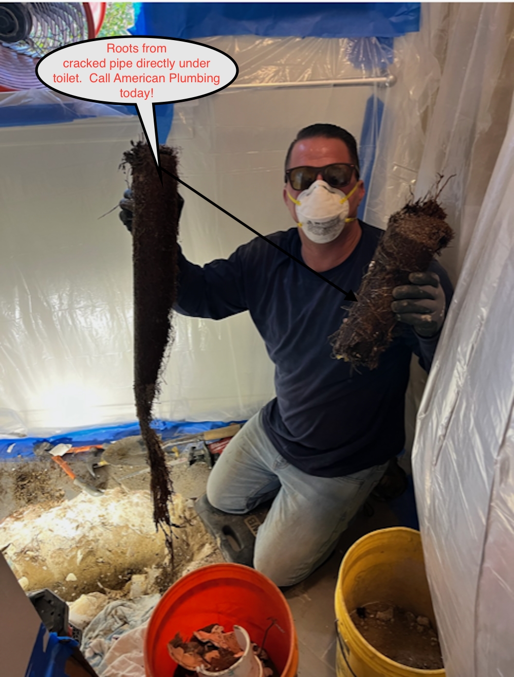 Worker in a plastic-covered room holds two clumps of insulation or debris over buckets, wearing a mask and sunglasses.