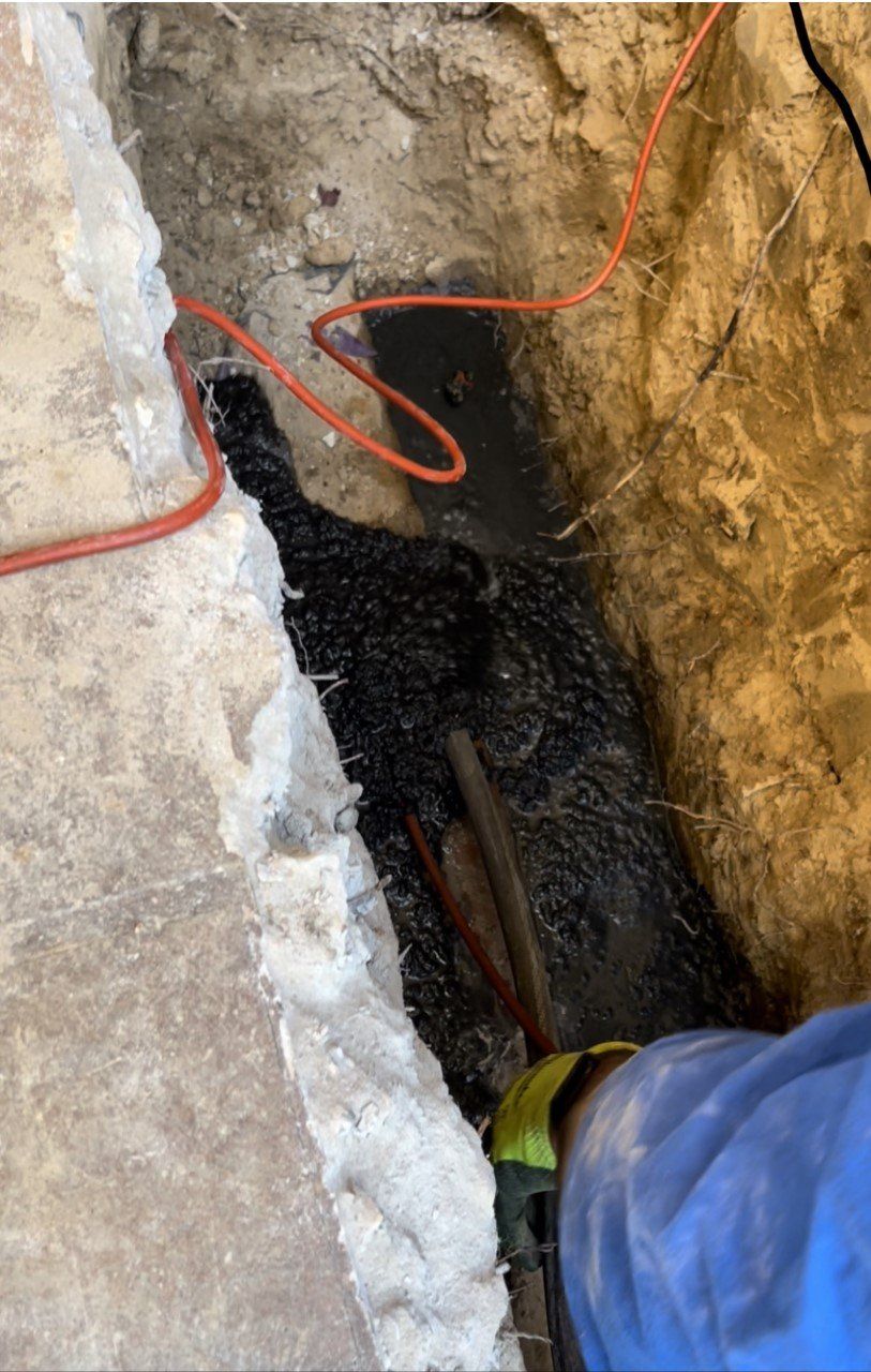 Person in a blue sleeve inspecting a narrow underground trench with exposed cables and pipes