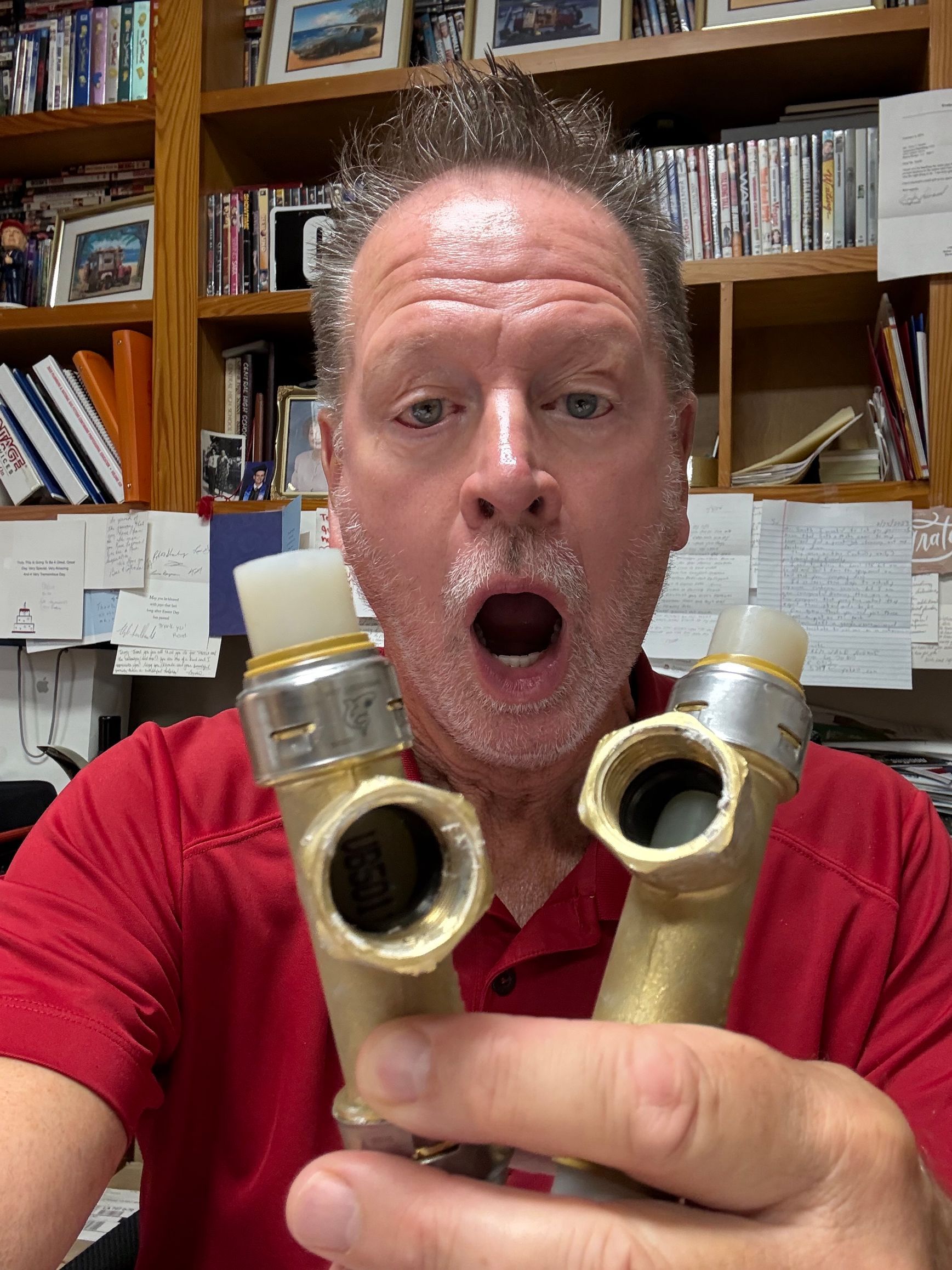 Man with surprised expression holds two brass plumbing fittings; office setting with bookshelves.