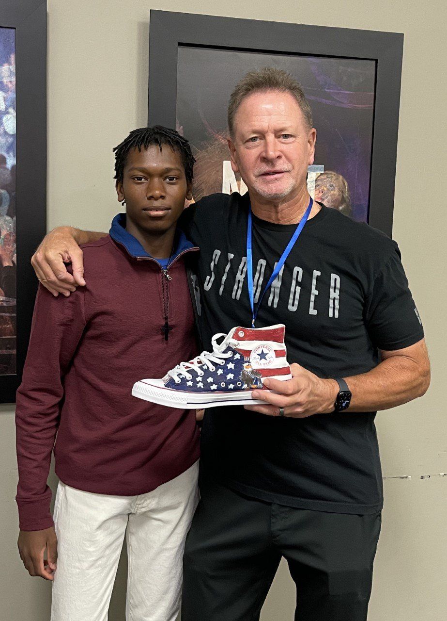 Man and teen holding American flag-painted sneaker, both smiling, indoors.