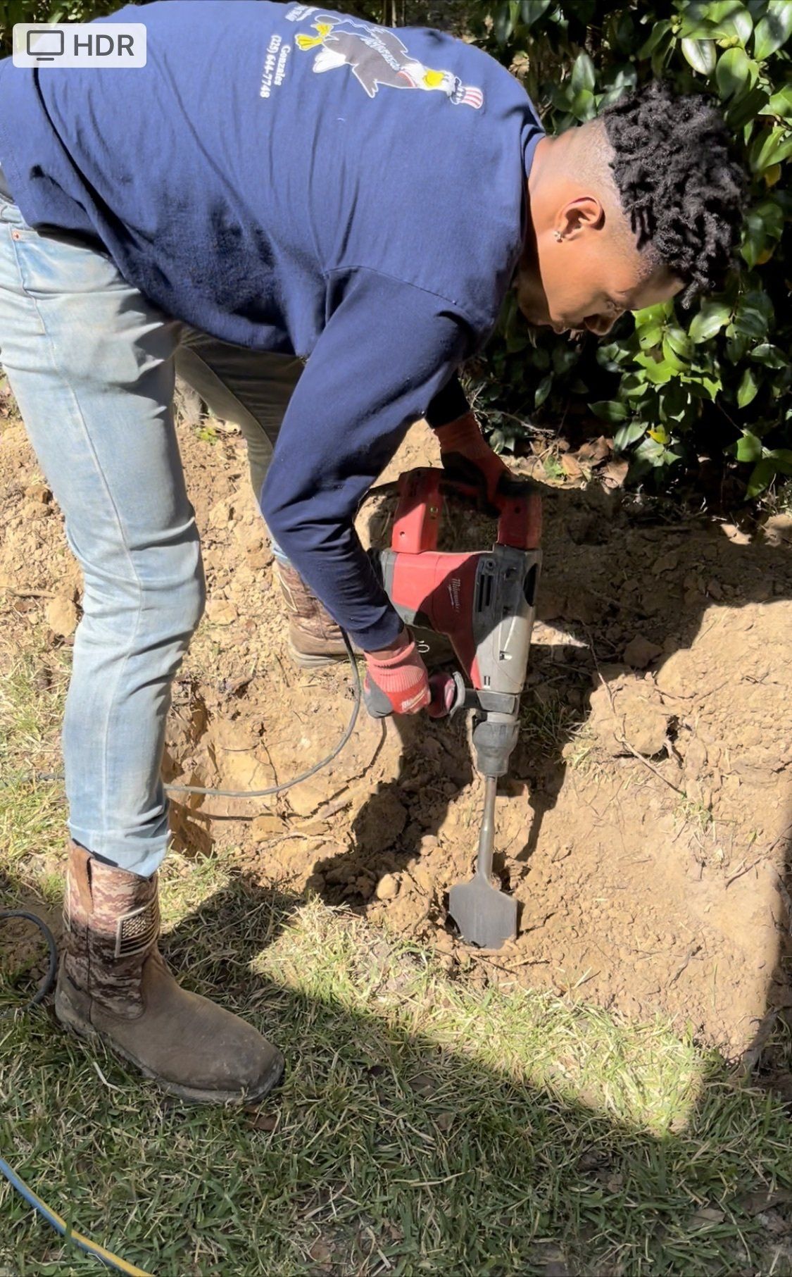 Man using a power drill on soil outdoors; wearing blue shirt, jeans, and brown boots.