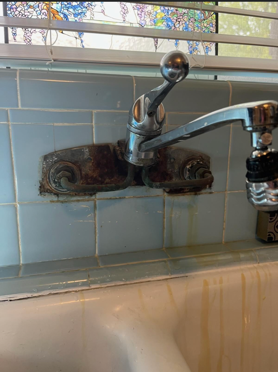 Rusty wall-mounted faucet above a white sink and blue tile backsplash under a window
