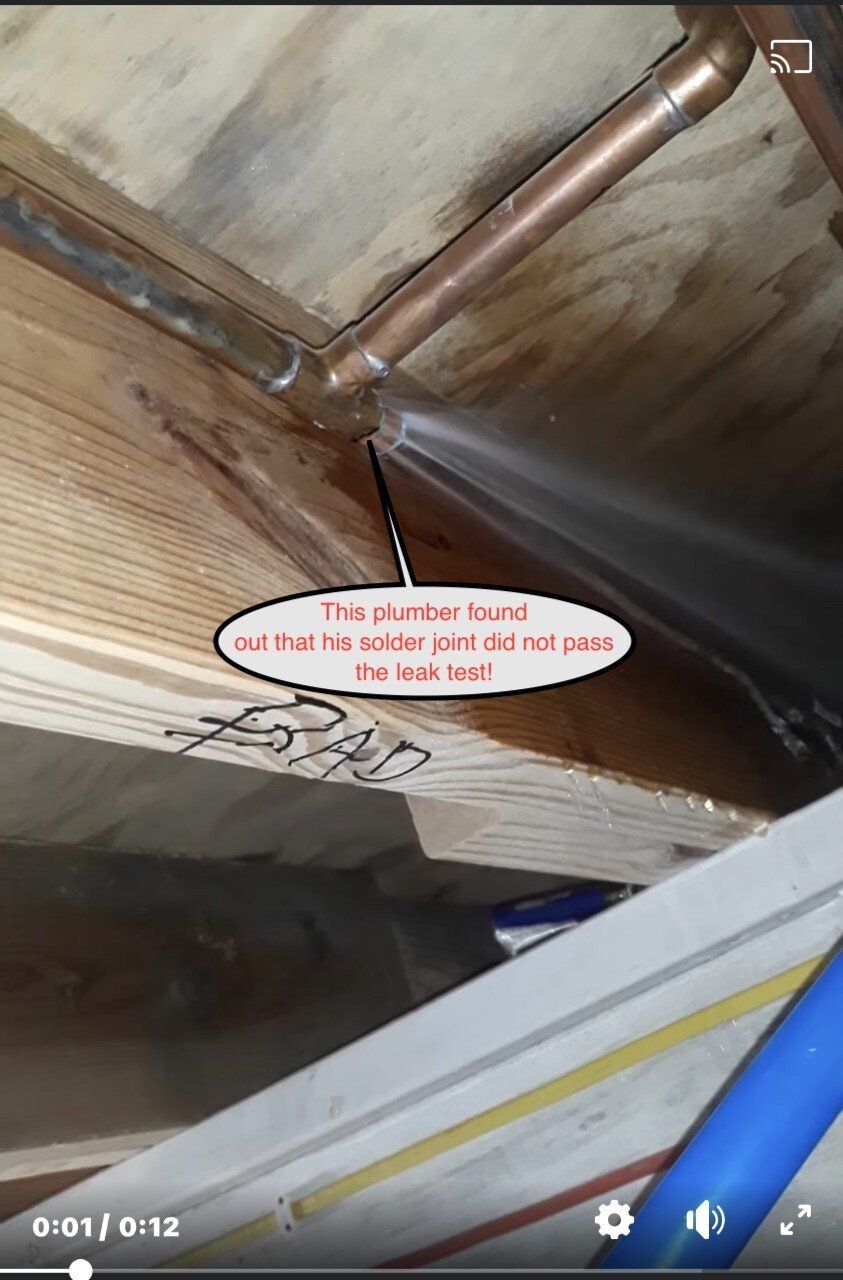 Water spraying from a copper pipe joint under a wood floor. Text on the image describes the leak.