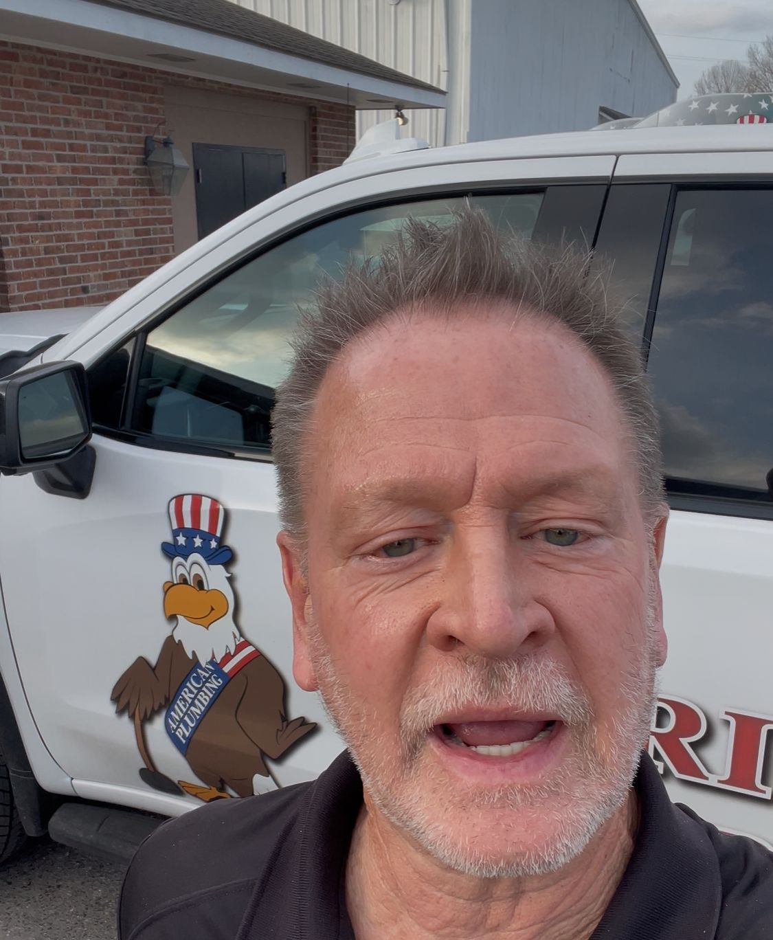 Man taking a selfie beside a white van with a cartoon mascot logo outdoors on a cloudy day