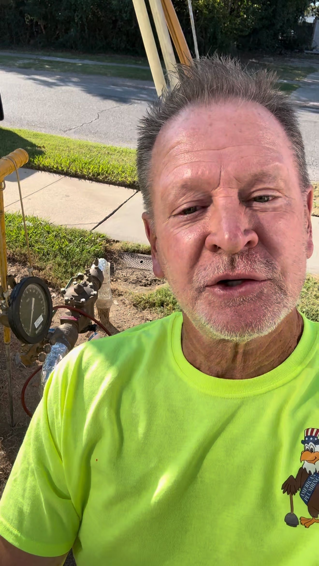 Man in neon yellow shirt taking a selfie outdoors on a sunny sidewalk near a street and grass.