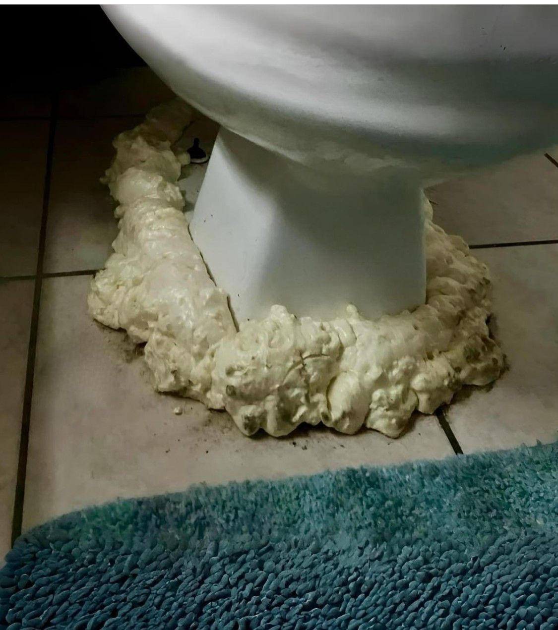 Toilet base surrounded by expanding white foam on a tiled bathroom floor near a blue rug