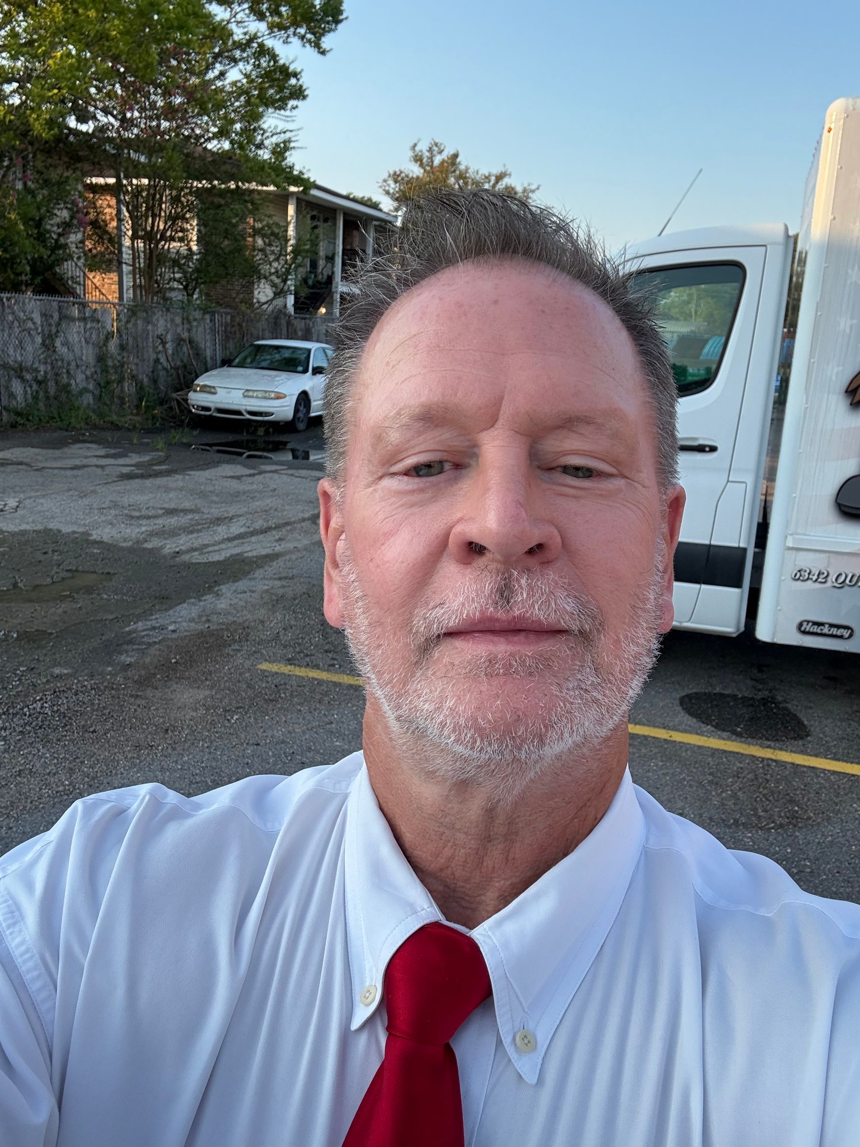 Man in white shirt and red tie taking a selfie outdoors near a truck and building at dusk