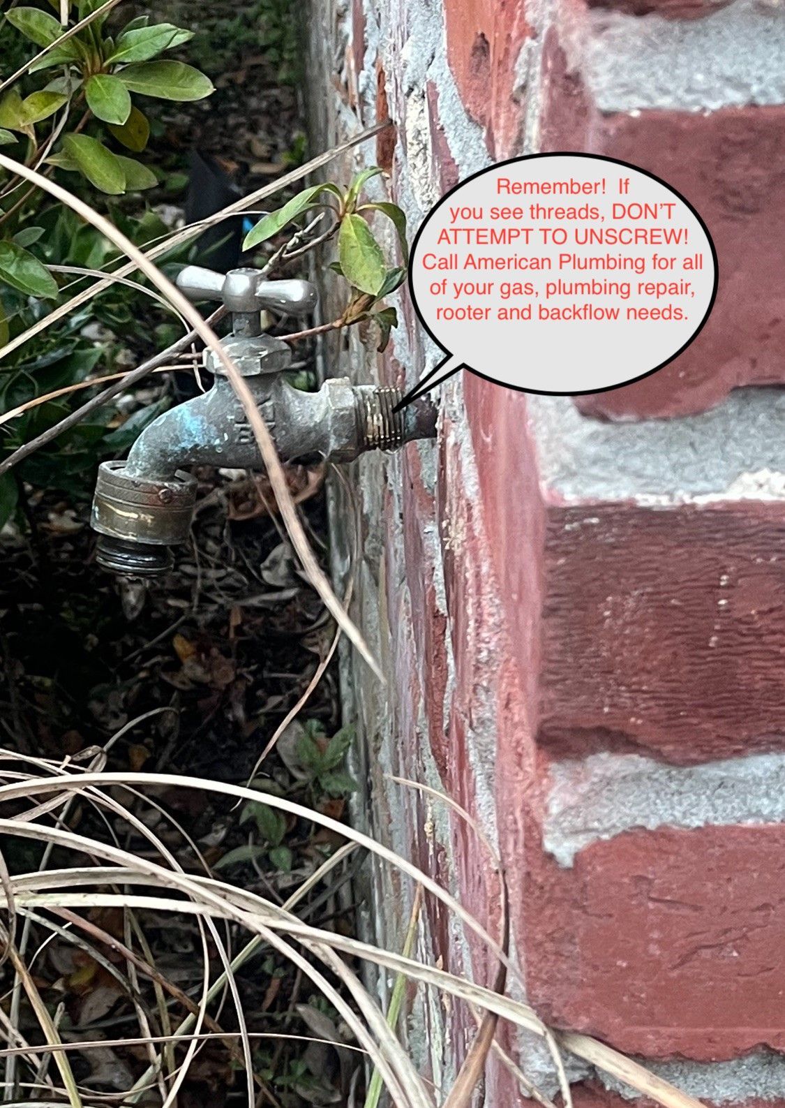 Faucet and hose beside a brick wall, partly obscured by dry grass and a warning sign.