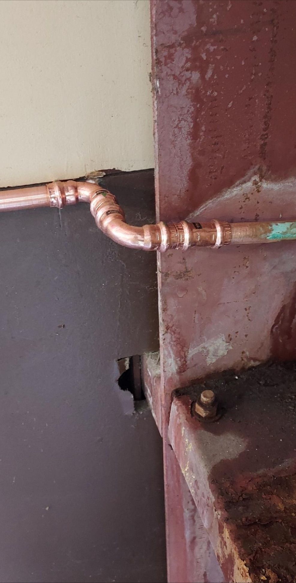 Copper pipe joint attached to a rusty metal surface near a wall opening.
