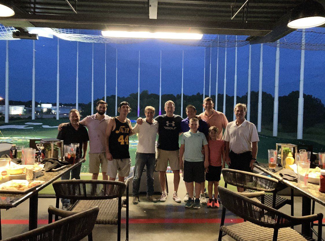 Group of people posing under a covered pavilion at dusk, with picnic tables and a sports field in the background