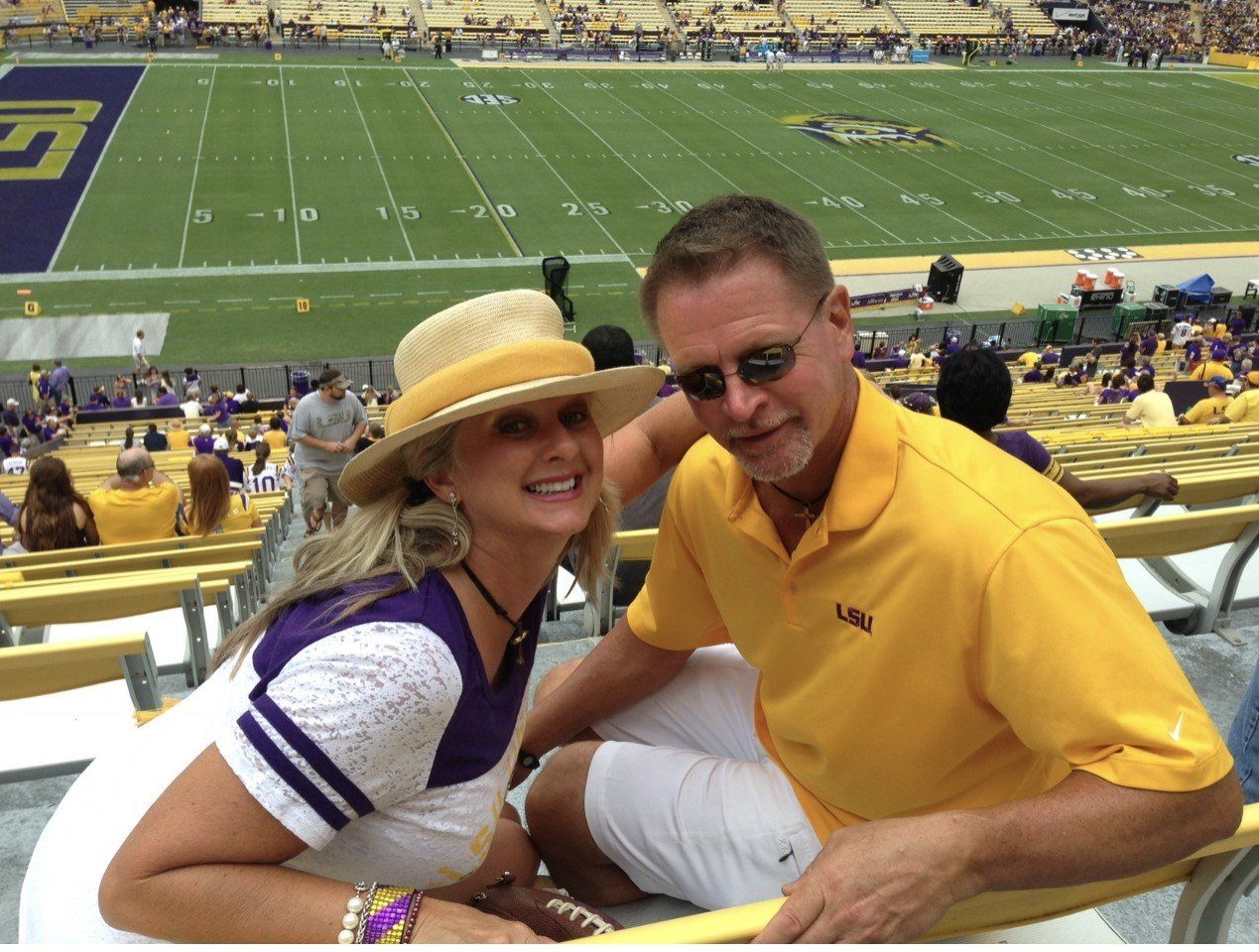 Two smiling fans in yellow and white sit at a football stadium with the field behind them.