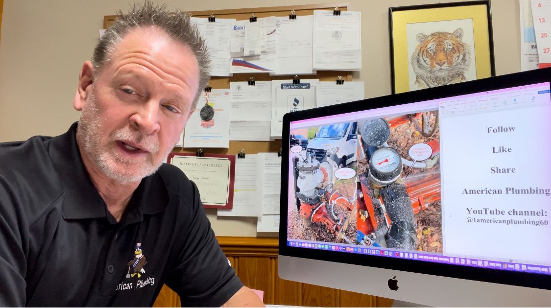 Man in black shirt speaking, pointing to computer screen with plumbing image. Background includes papers and tiger art.