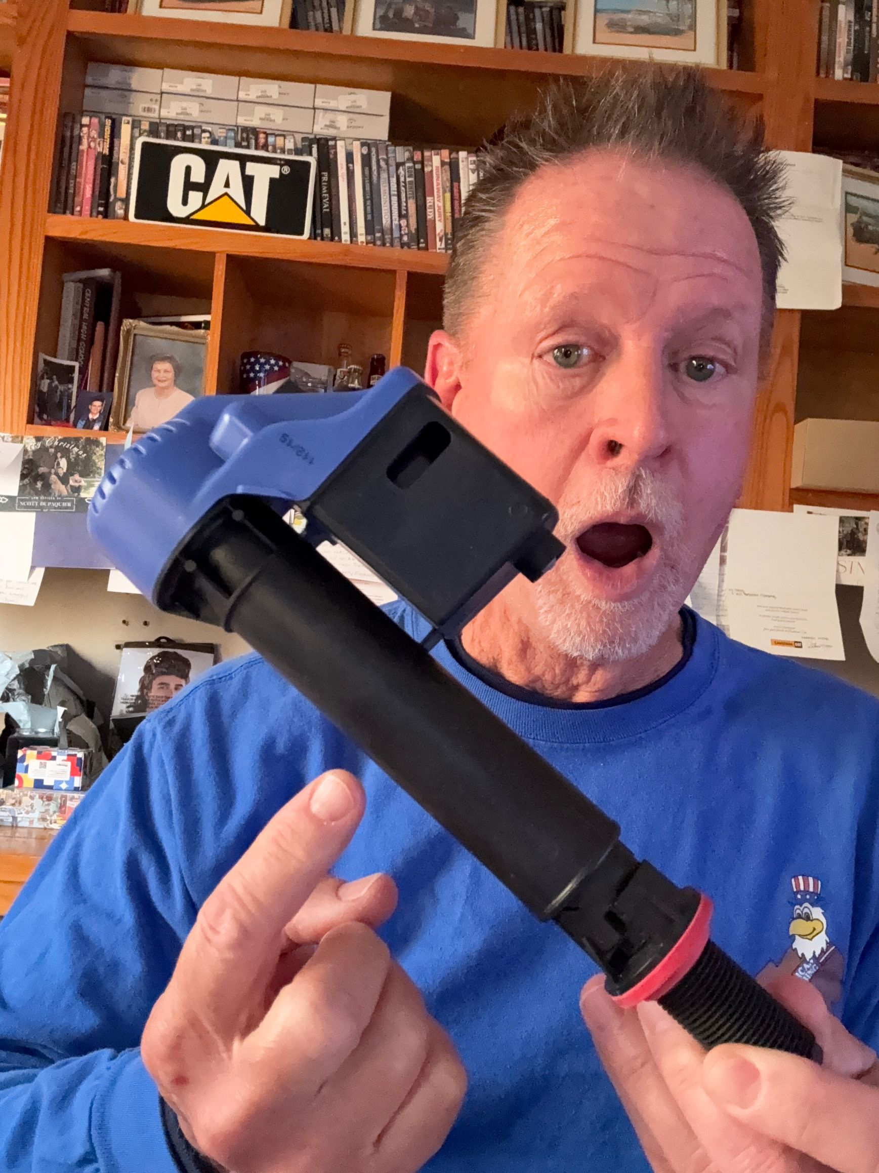 Man holding a black-and-blue tool, mouth open in surprise, in a cluttered home workshop