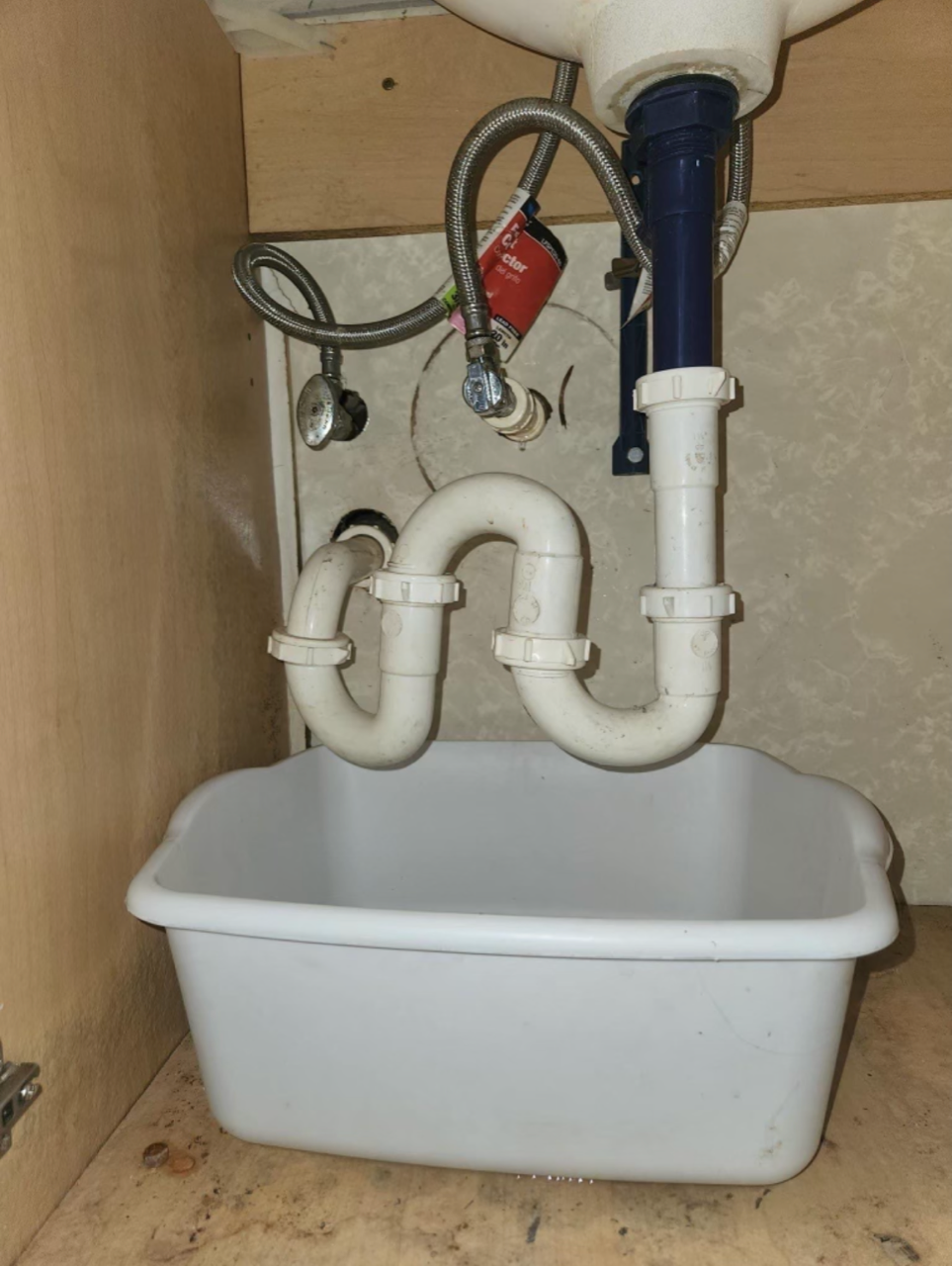 White sink plumbing under a sink, with a bucket underneath to catch potential leaks.