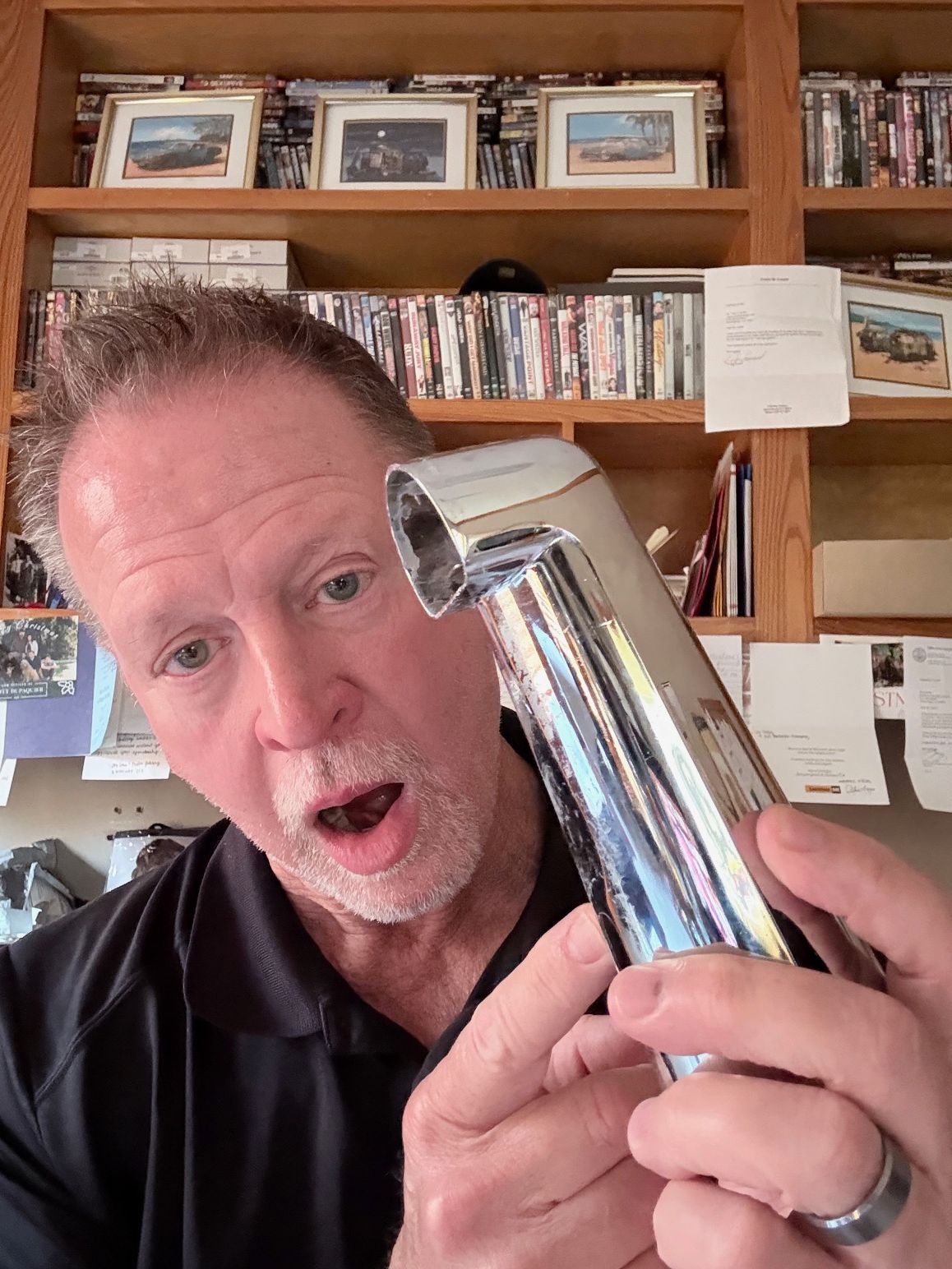 Man holding a shiny chrome object close to his face in a home office with shelves and framed photos behind him