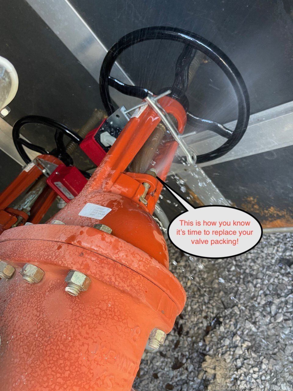 Red industrial valve with black handwheel and orange housing in a concrete utility area.