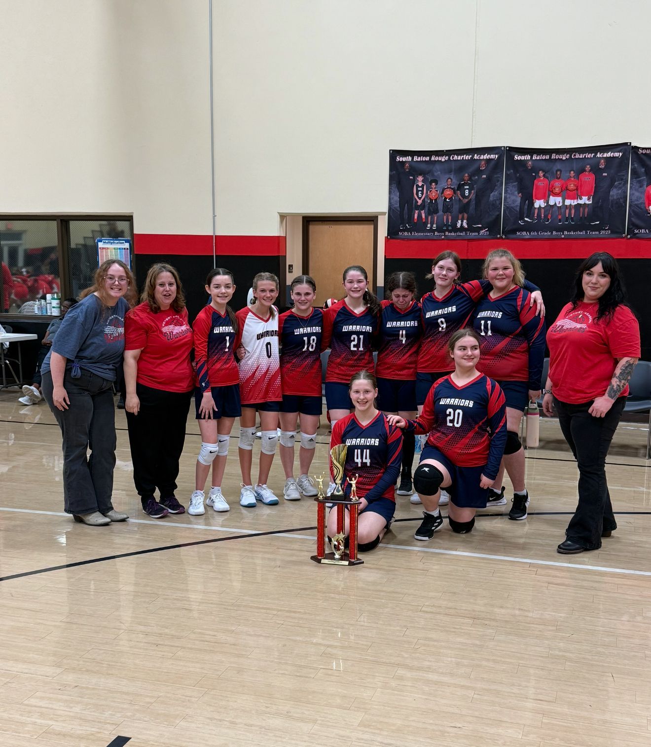 Youth volleyball team in red and blue uniforms, posing with a trophy in a gym.