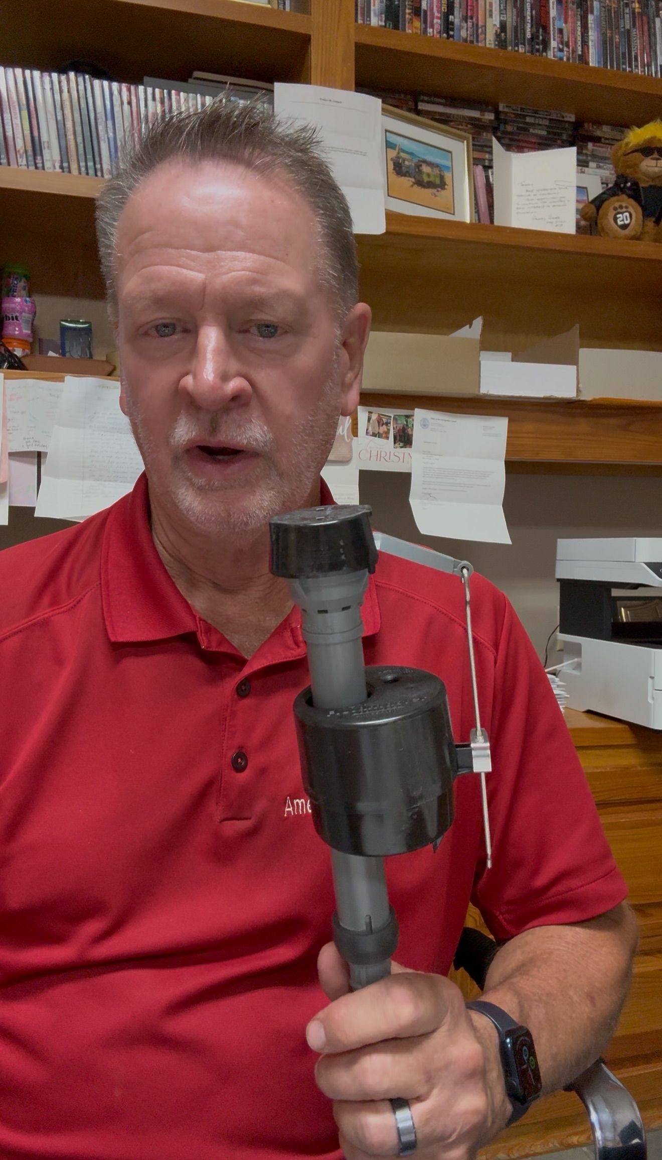 Man in red shirt holds a toilet fill valve, explaining its parts. Brown bookshelf background.