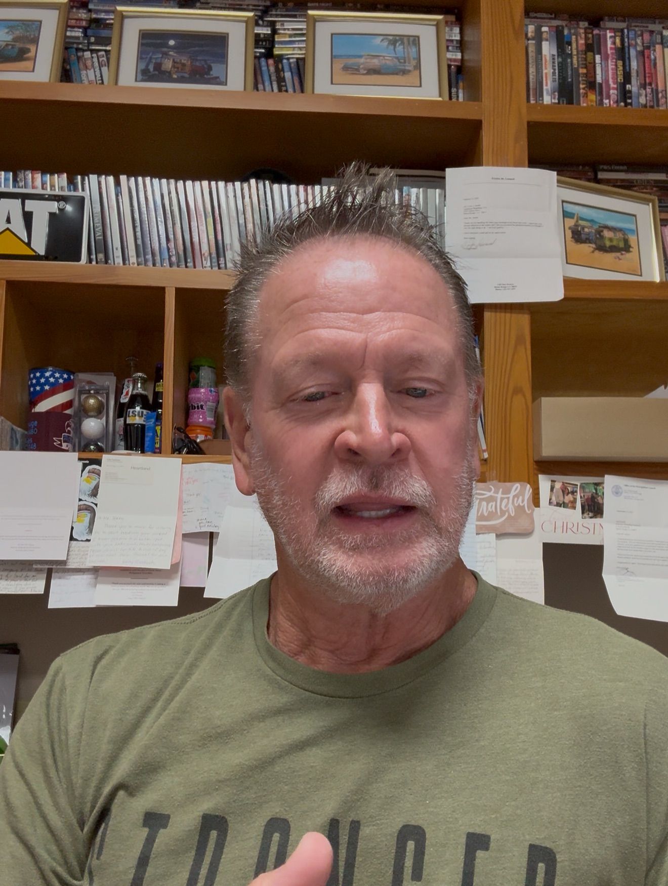 Man in olive green shirt speaking; bookshelf background.
