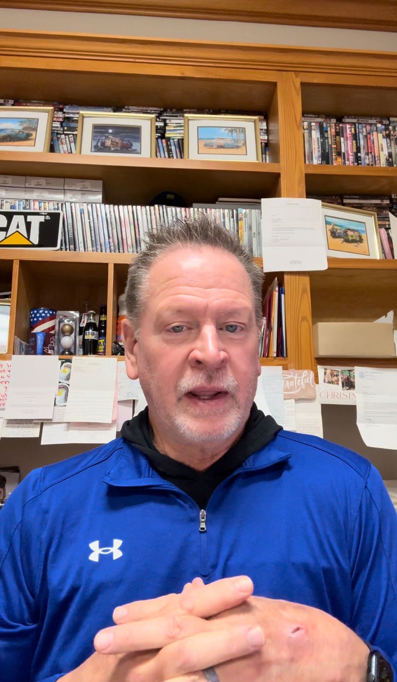 Man in blue quarter-zip speaking in front of wooden shelves with books and framed photos